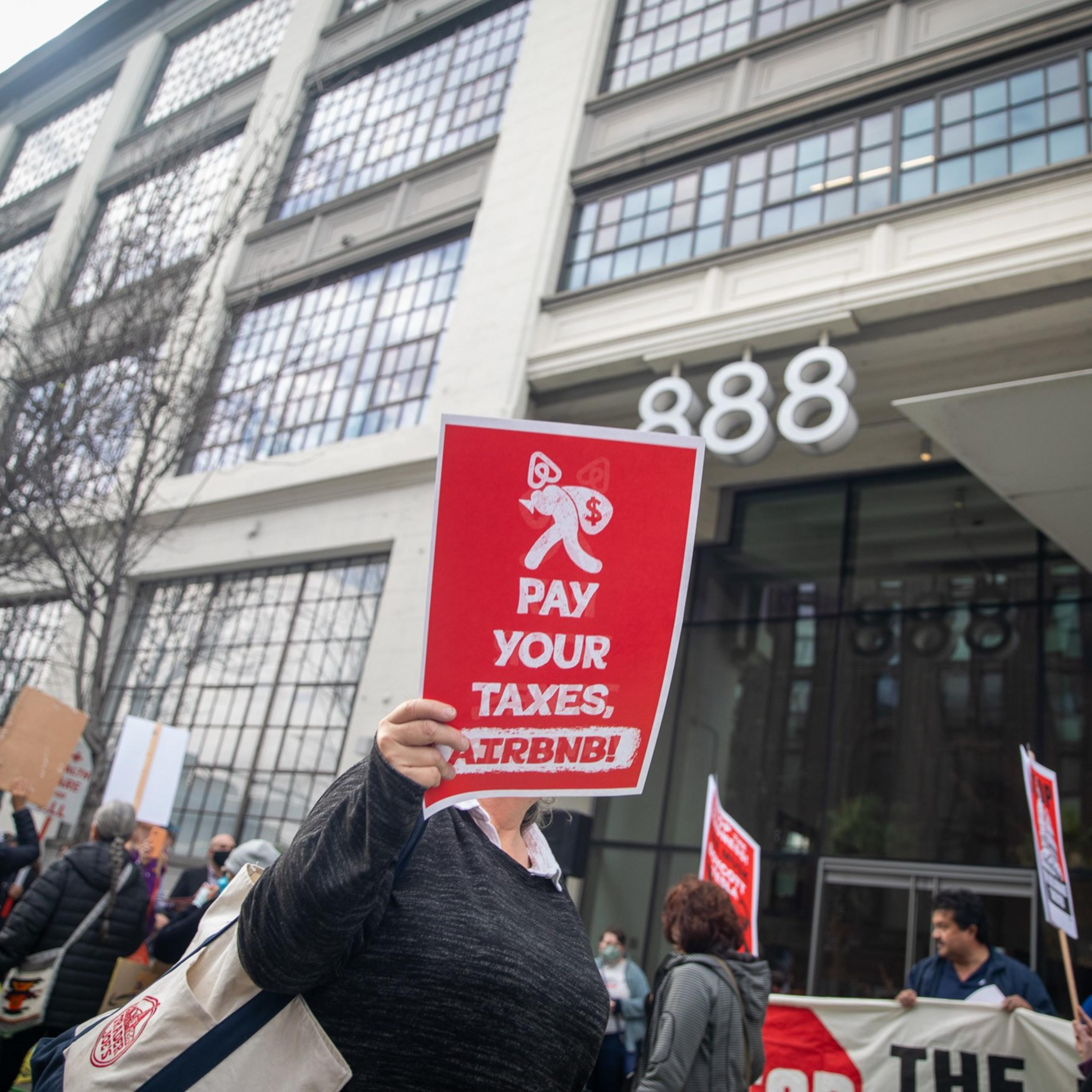 Protesters stand outside a large building, holding signs that say "PAY YOUR TAXES, AIRBNB!" The building has large windows and the number 888 visible.