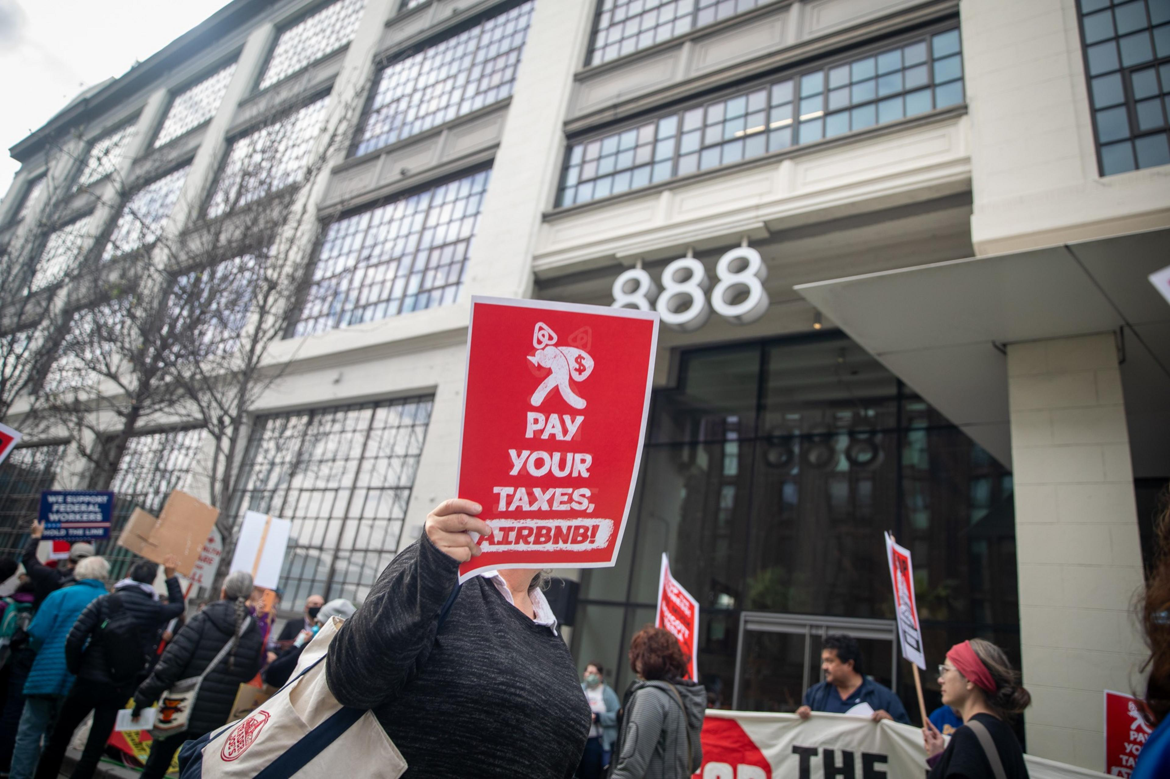 Protesters stand outside a large building, holding signs that say "PAY YOUR TAXES, AIRBNB!" The building has large windows and the number 888 visible.