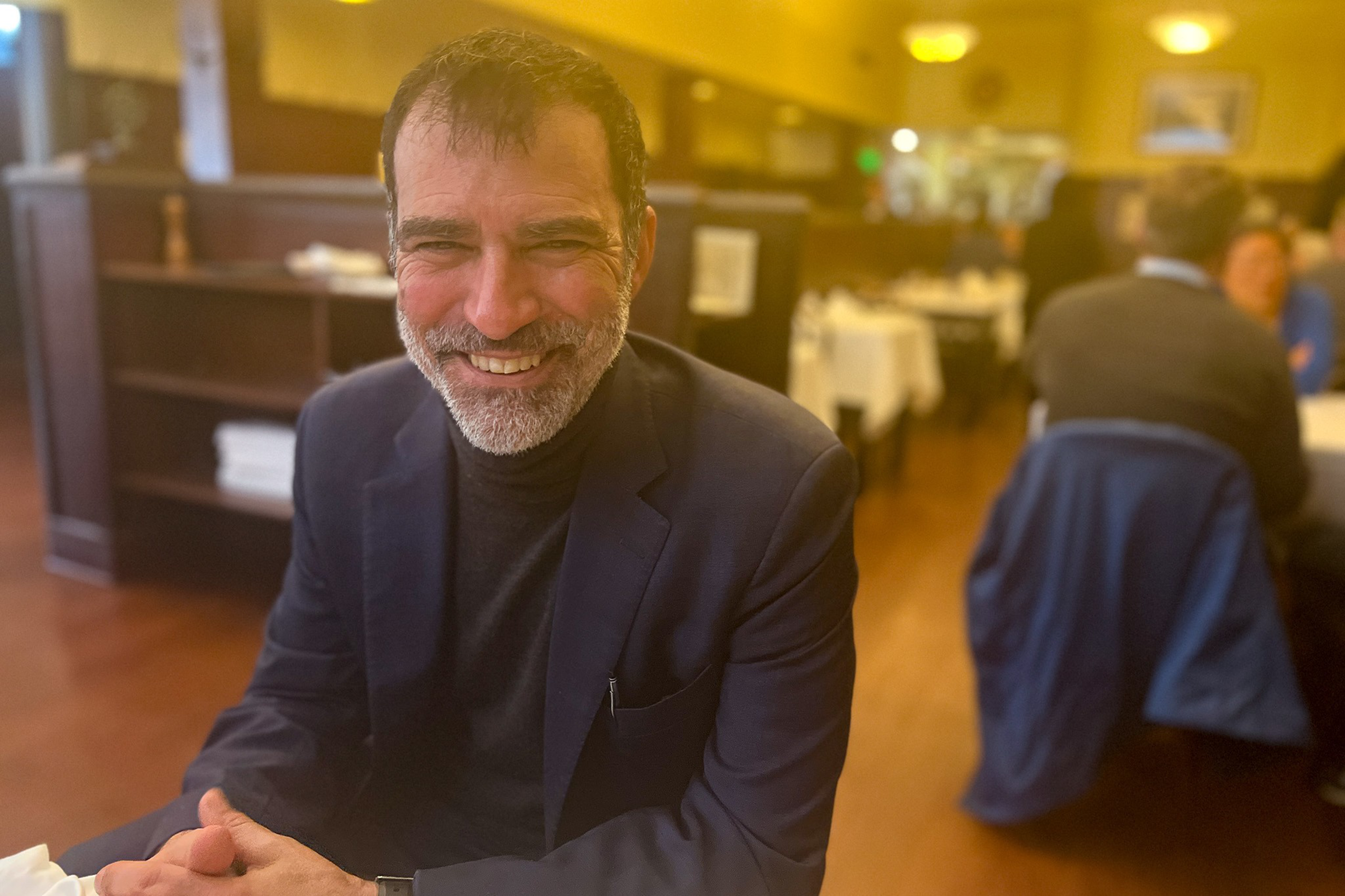 A man with short dark hair and a beard smiles warmly while sitting at a table in a warmly lit restaurant wearing a dark jacket and turtleneck.