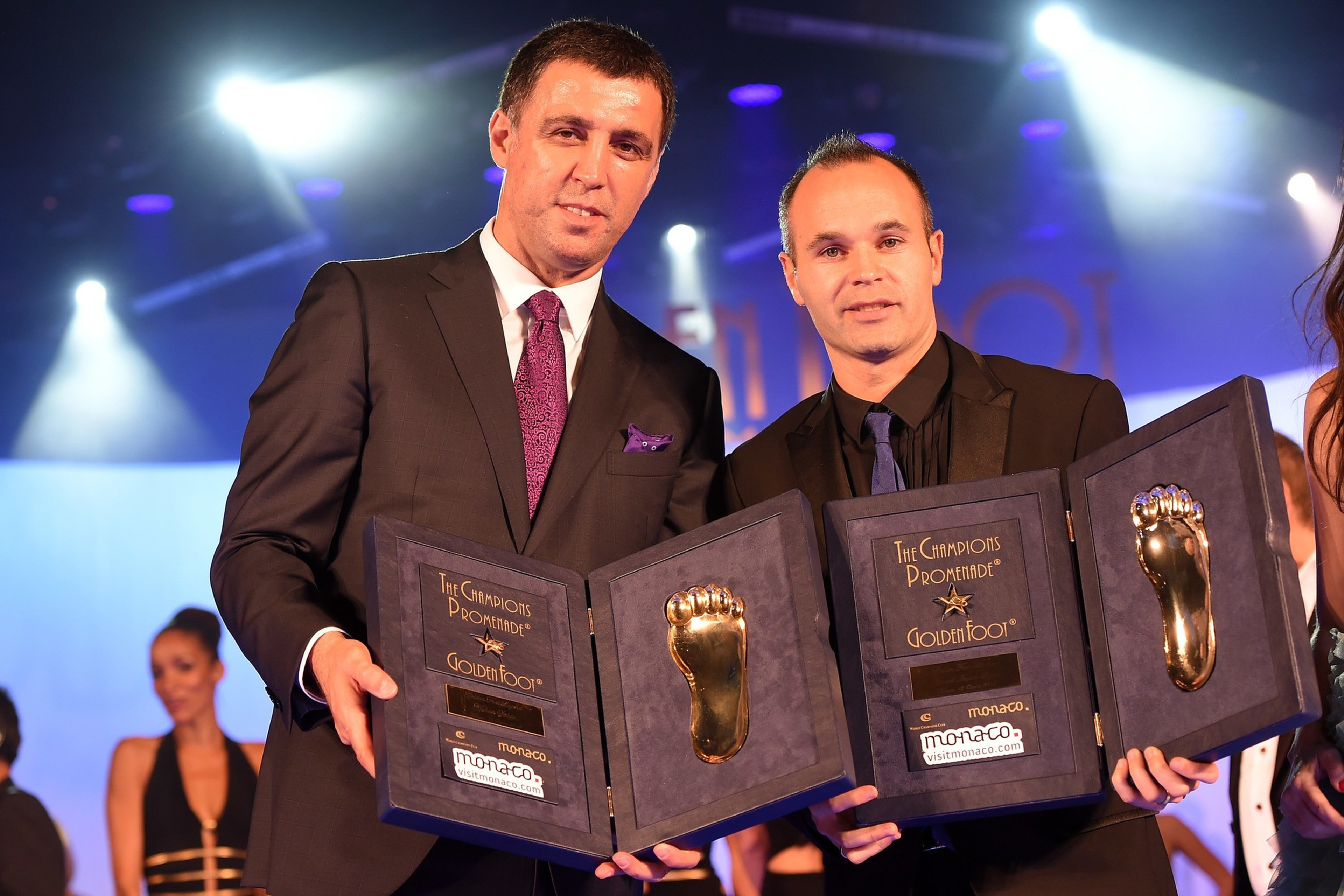 Two men in suits hold open plaques with golden foot trophies, standing on a stage with bright lights and blurred people in the background.