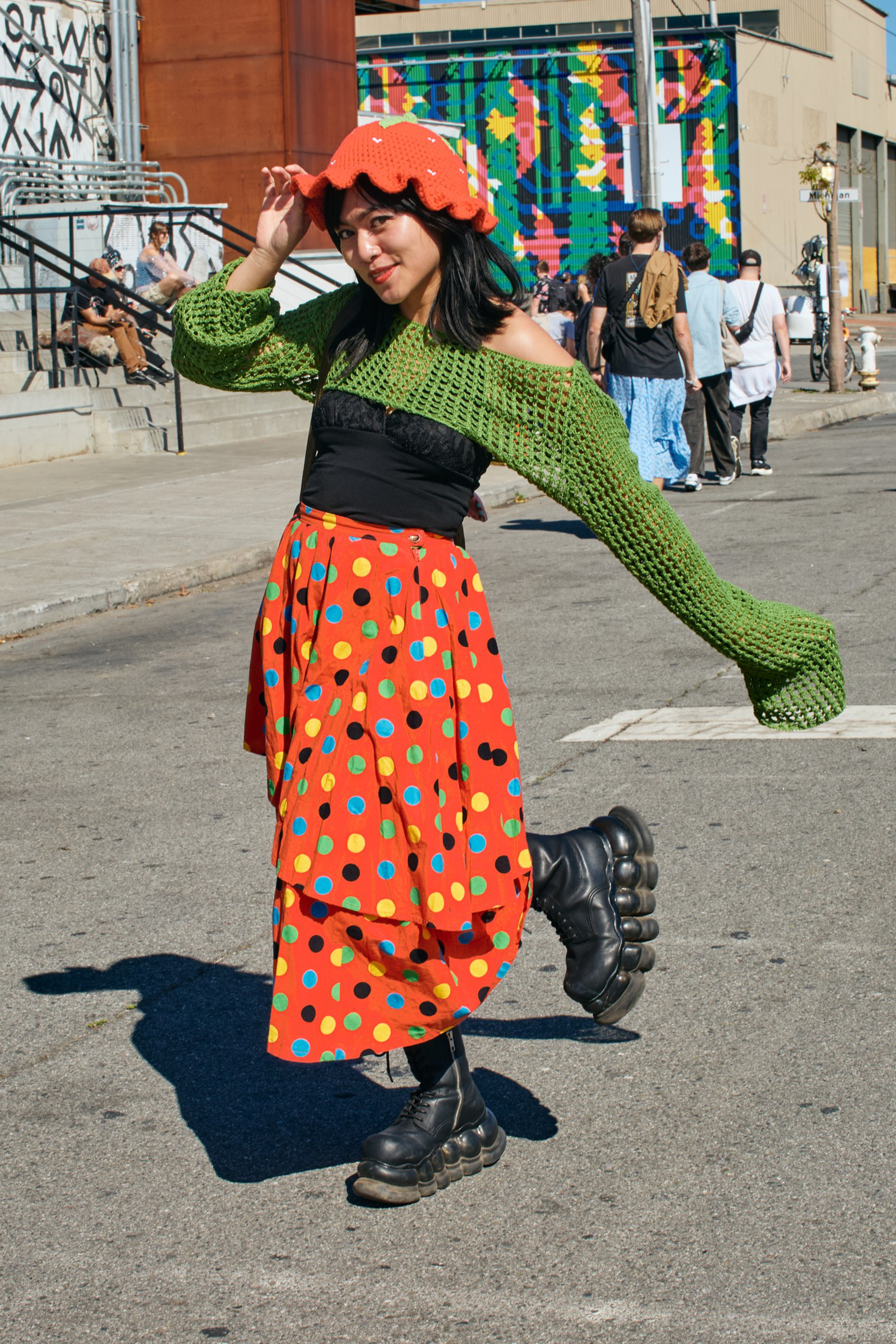 A young woman wearing a red polka-dot skirt, green knitted off-shoulder sweater, red hat, and chunky black boots poses playfully on a sunny street.