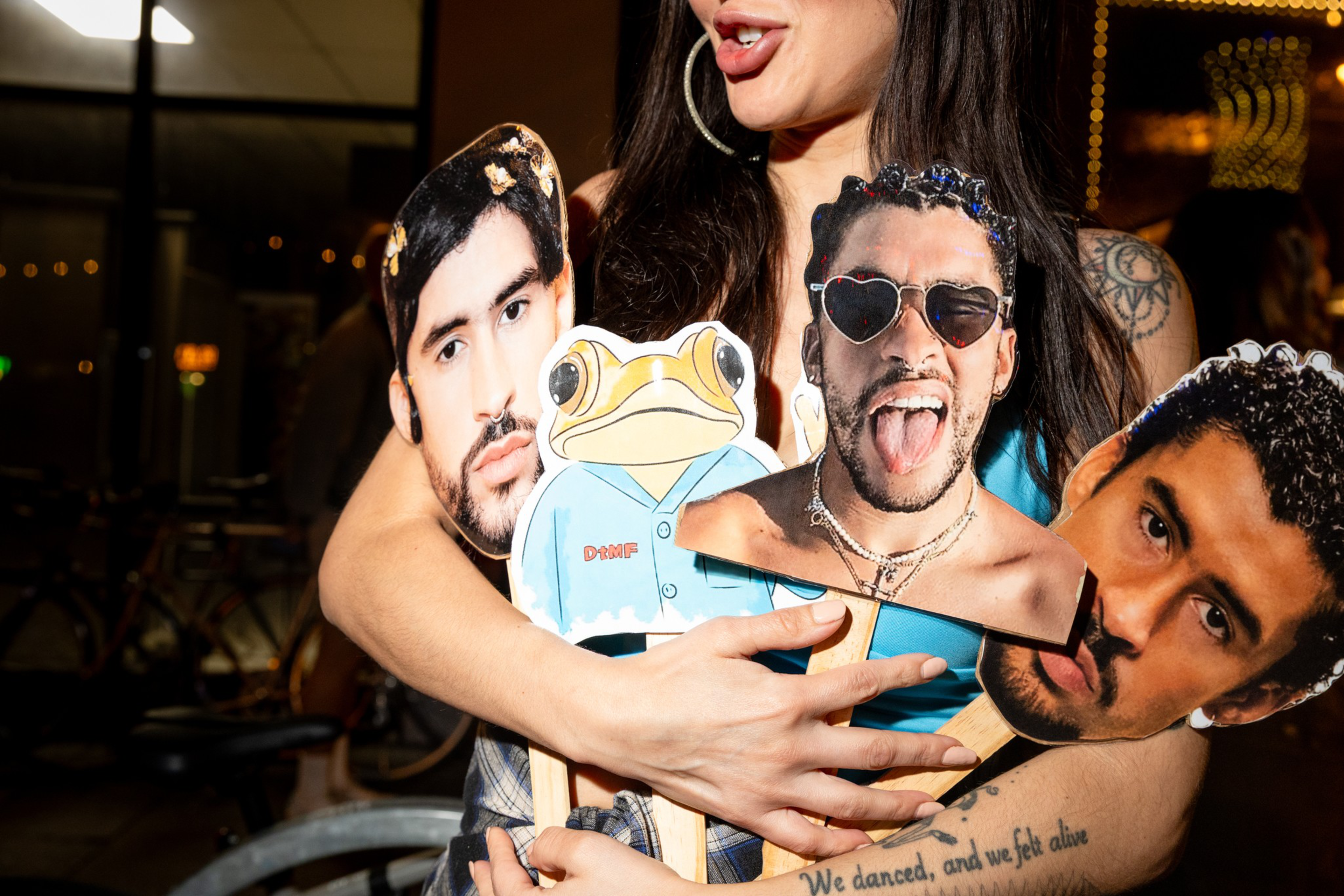 A person holds four cardboard cutouts on sticks: three male faces and one cartoon frog wearing a blue shirt.