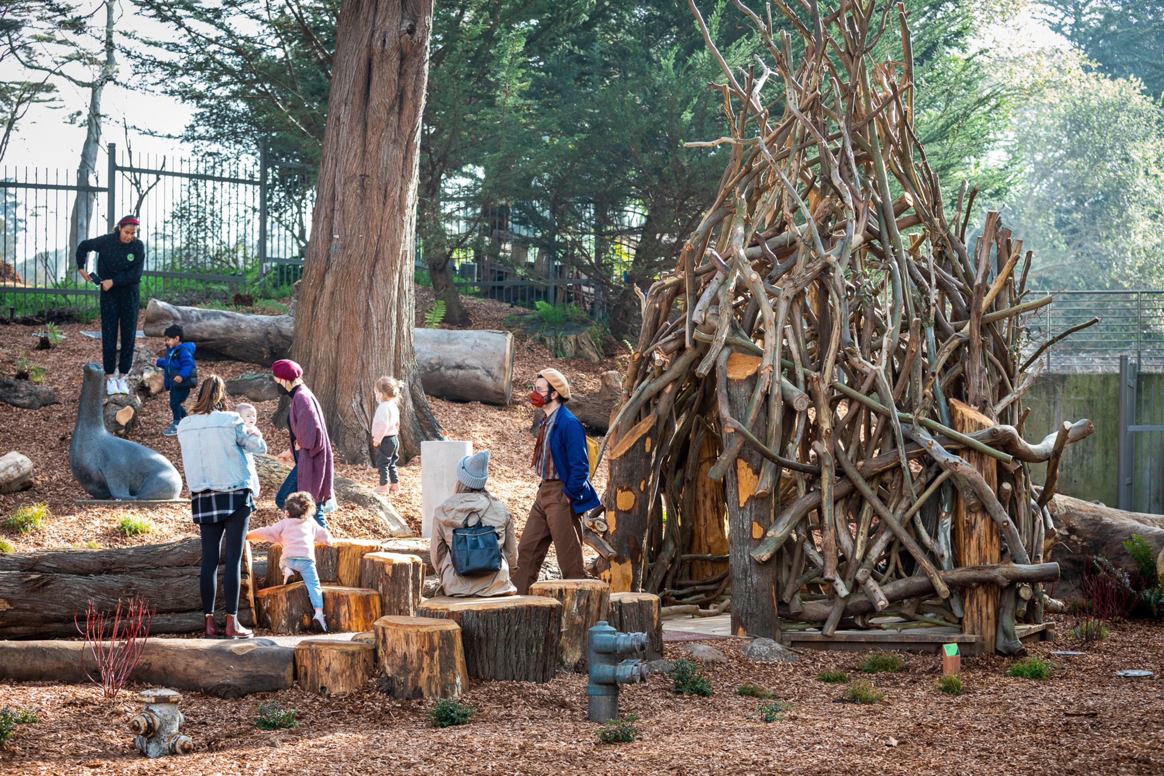 Children and adults play on wooden logs and climb a sculpture shaped like a seal near a teepee-like structure made of intertwined branches.