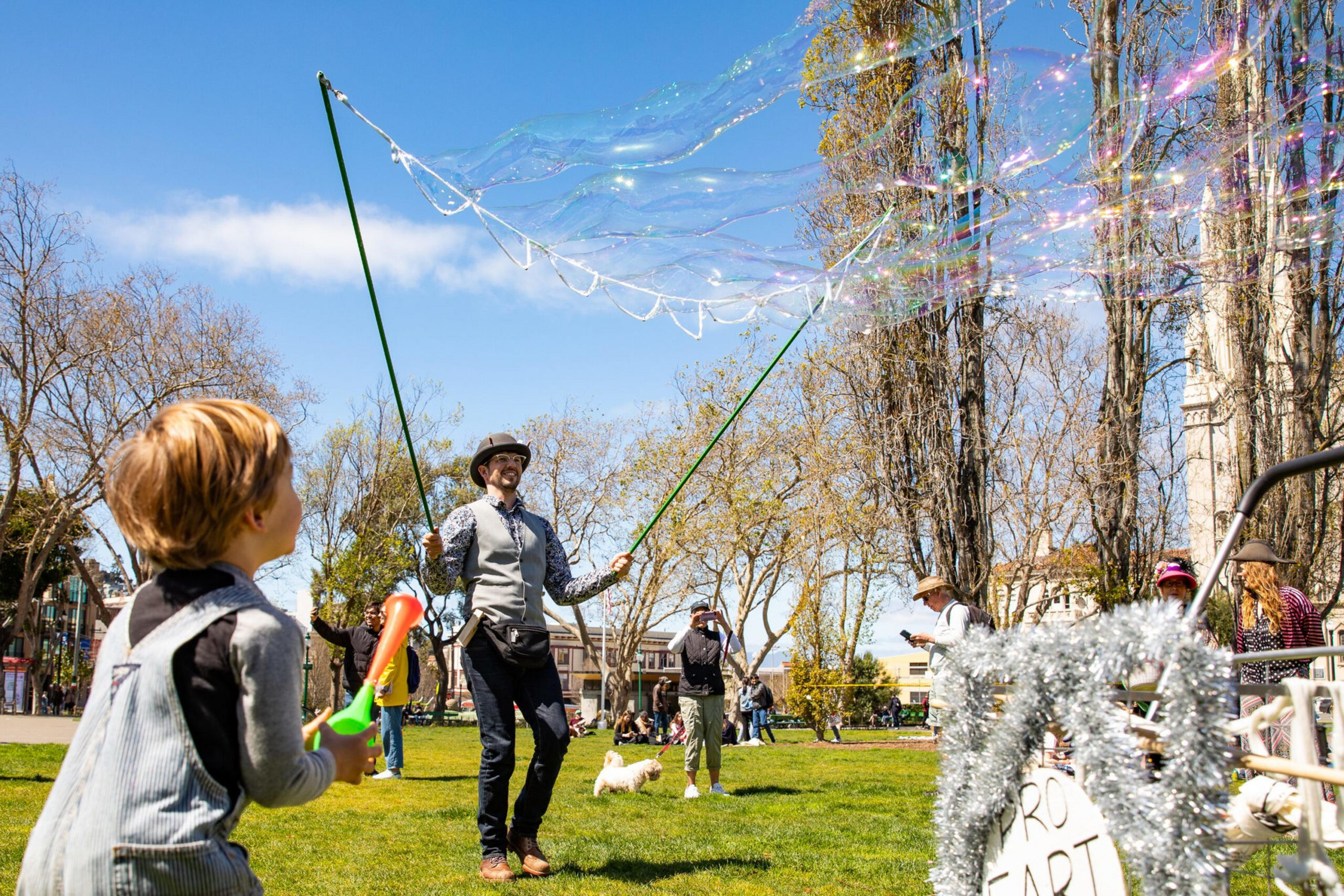 A man in a vest and hat creates giant bubbles with two sticks while a child watches and holds a bubble wand in a sunny park.