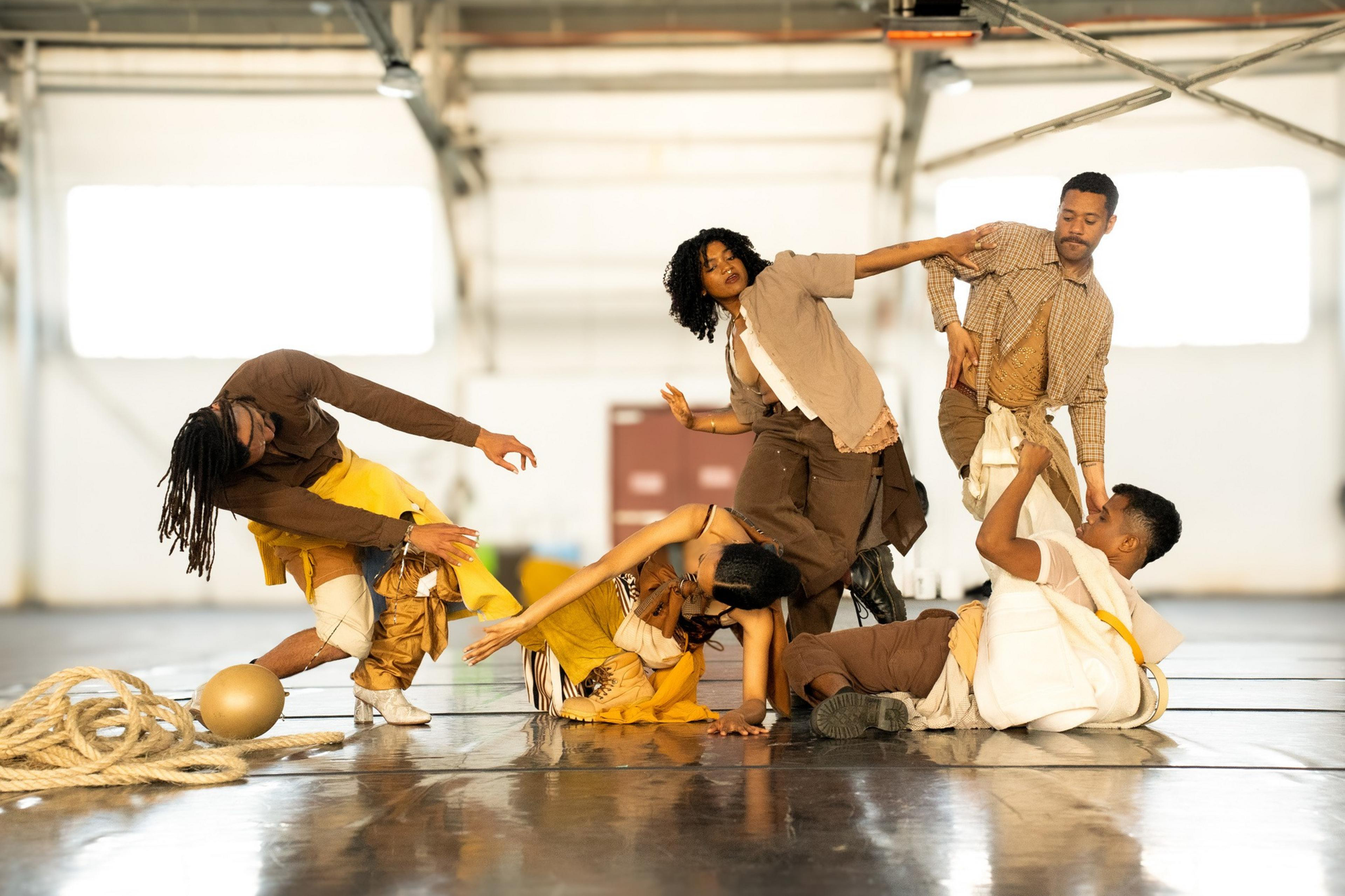 Five dancers in earthy tones perform expressive, intertwined movements on a shiny floor inside a spacious, light-filled industrial space.