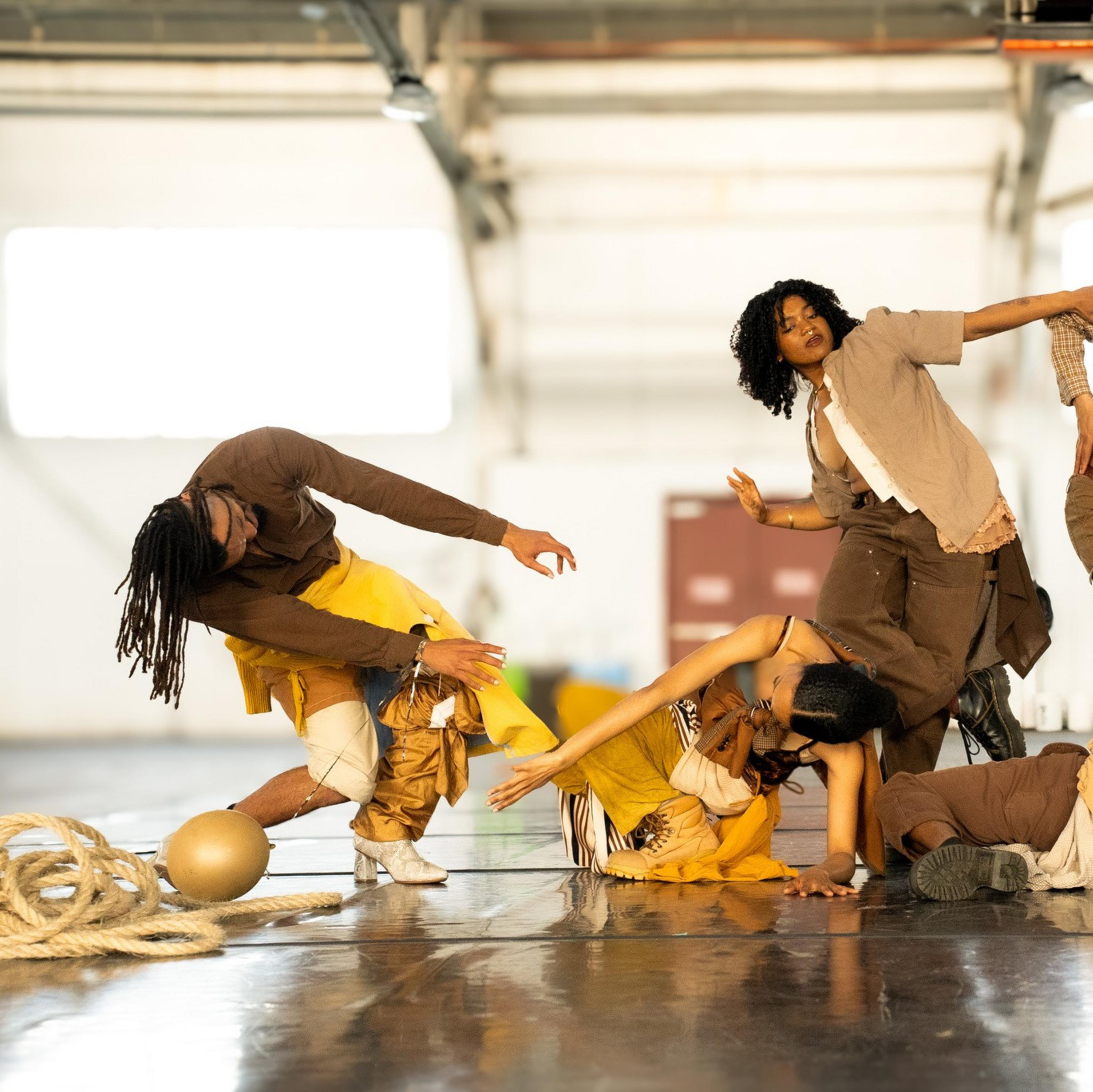 Five dancers in earthy tones perform expressive, intertwined movements on a shiny floor inside a spacious, light-filled industrial space.