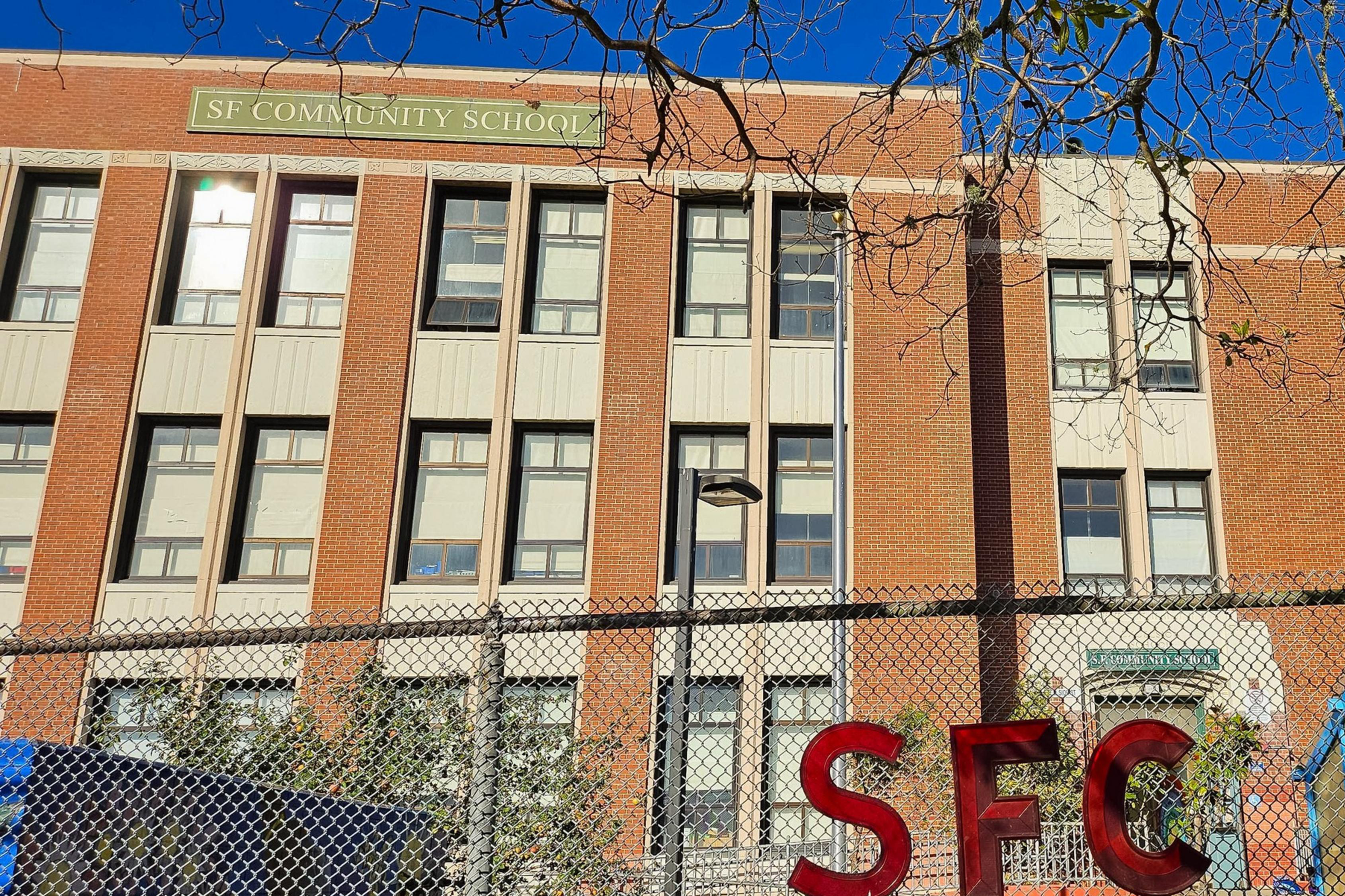 A brick building with a "SF Community School" sign is visible. In front, a chain-link fence features red letters spelling "SFC." Trees and a bright blue sky surround it.