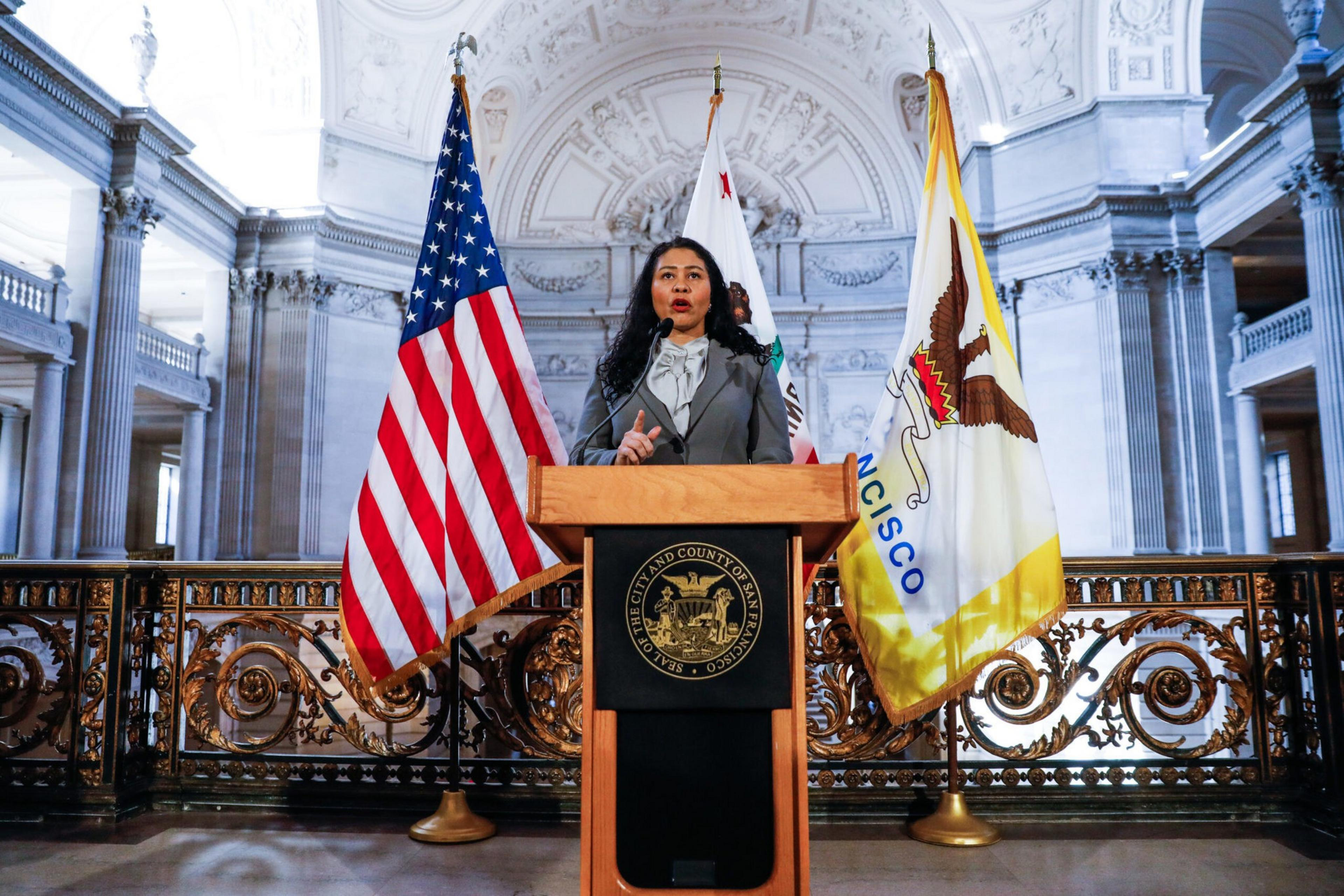 A woman in formal attire speaks at a wooden podium with San Francisco and U.S. flags behind her in an ornate government building.