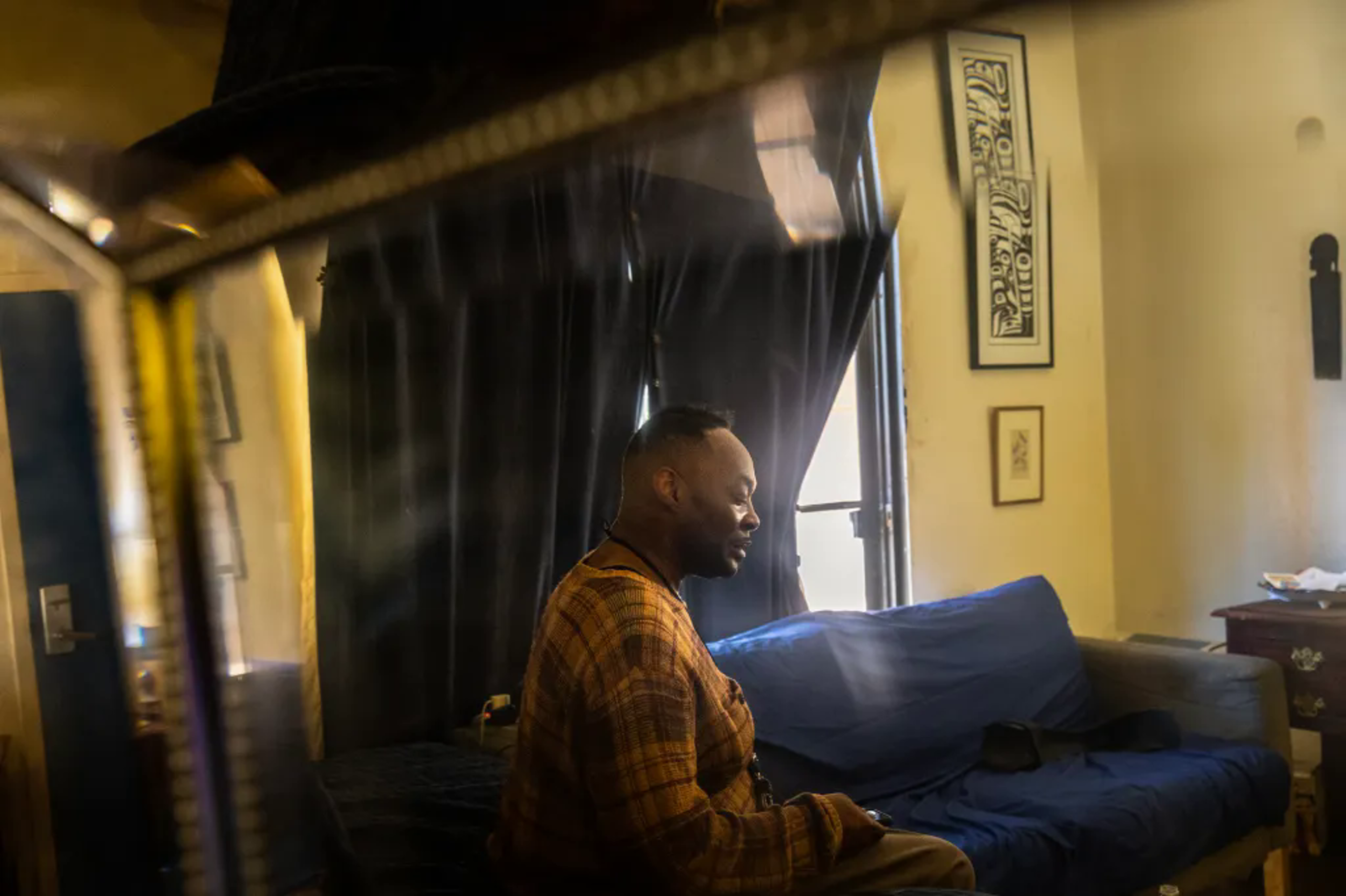 Man sits inside apartment with dark curtains and couch covered with blue sheet