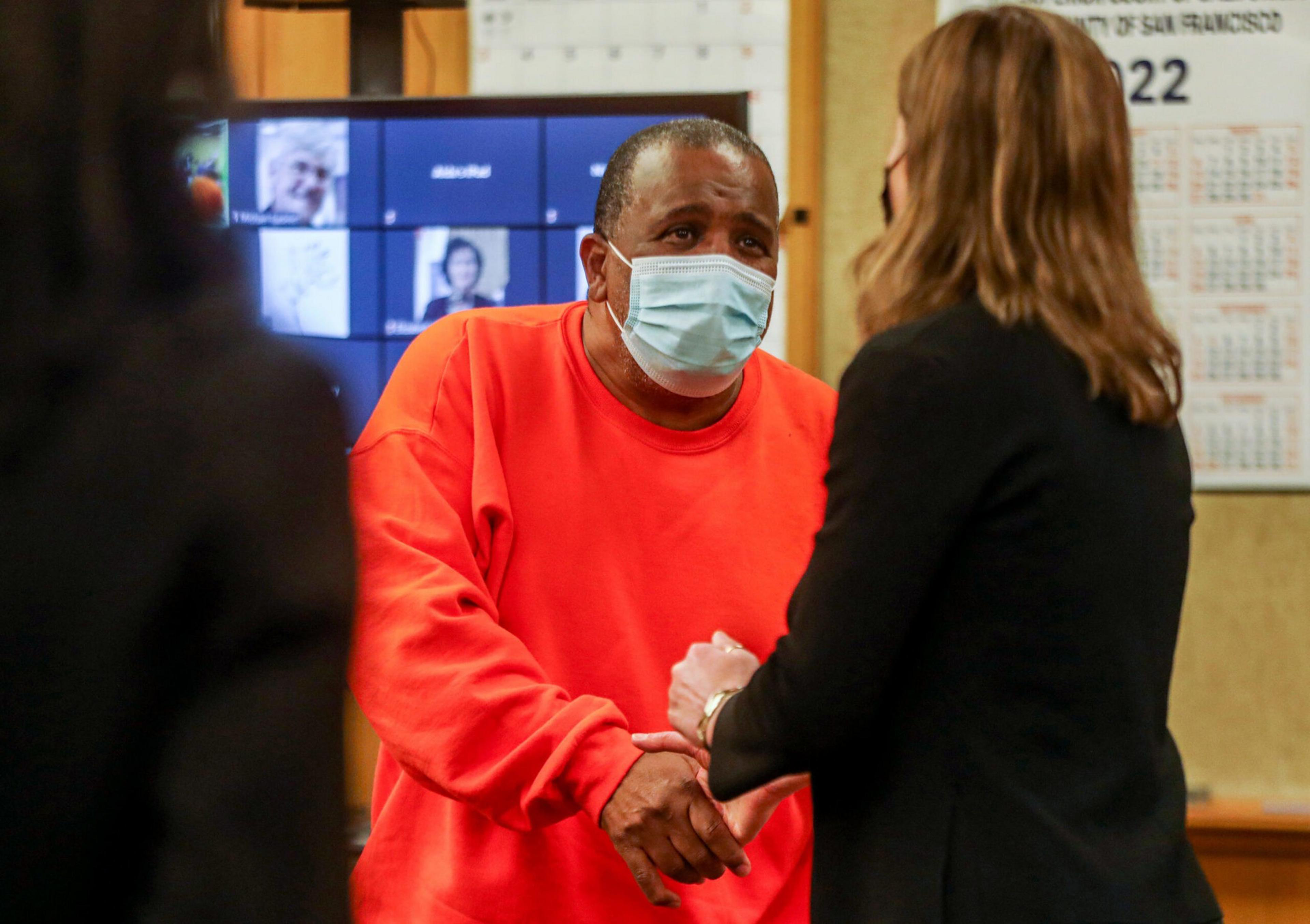 A man in a bright orange shirt and face mask shakes hands with a woman wearing a black jacket in an indoor setting with screens and calendars in the background.