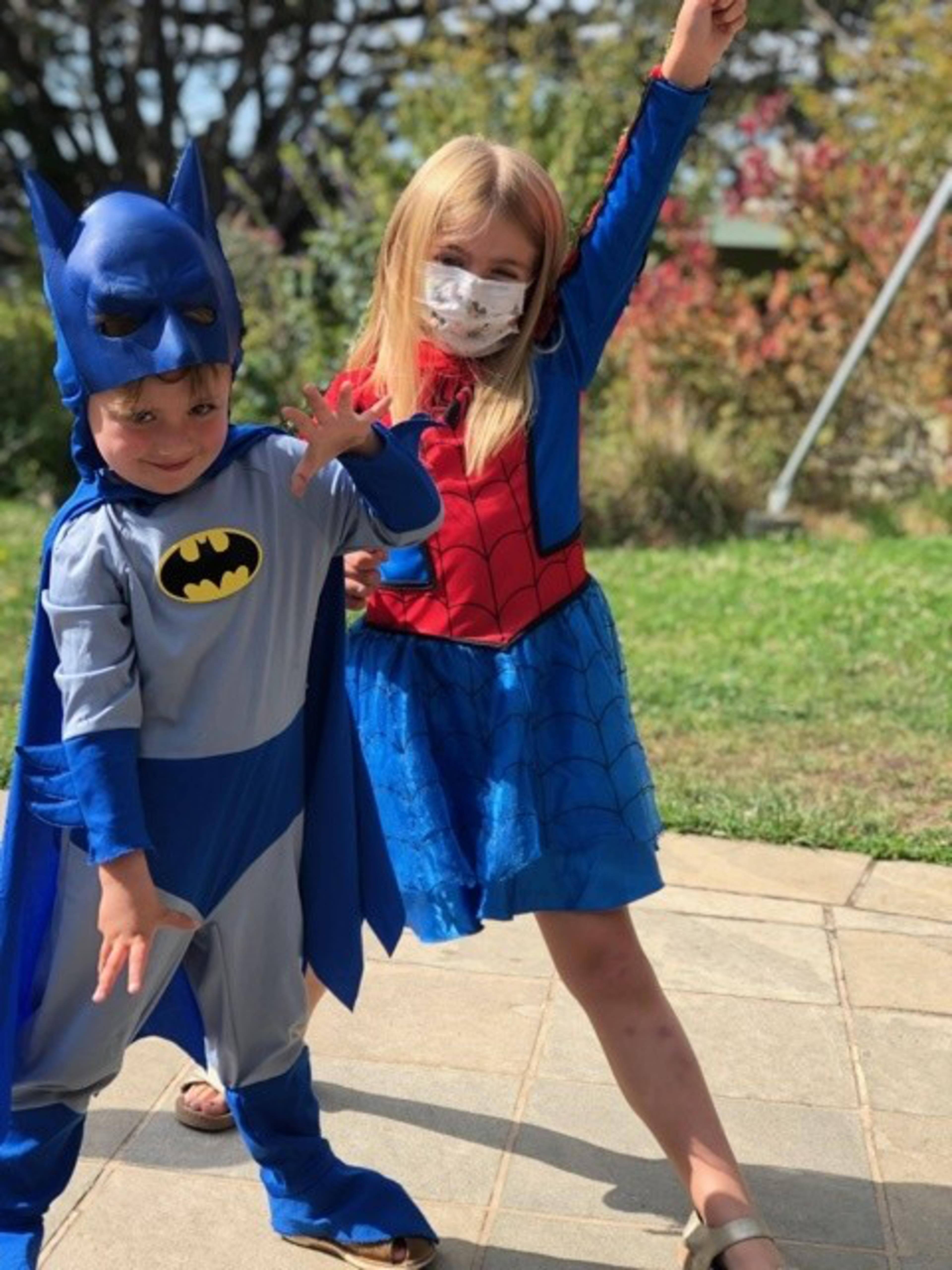 A boy in a Batman costume and a girl in a Spider-Man dress and mask pose playfully outdoors on a sunny day.
