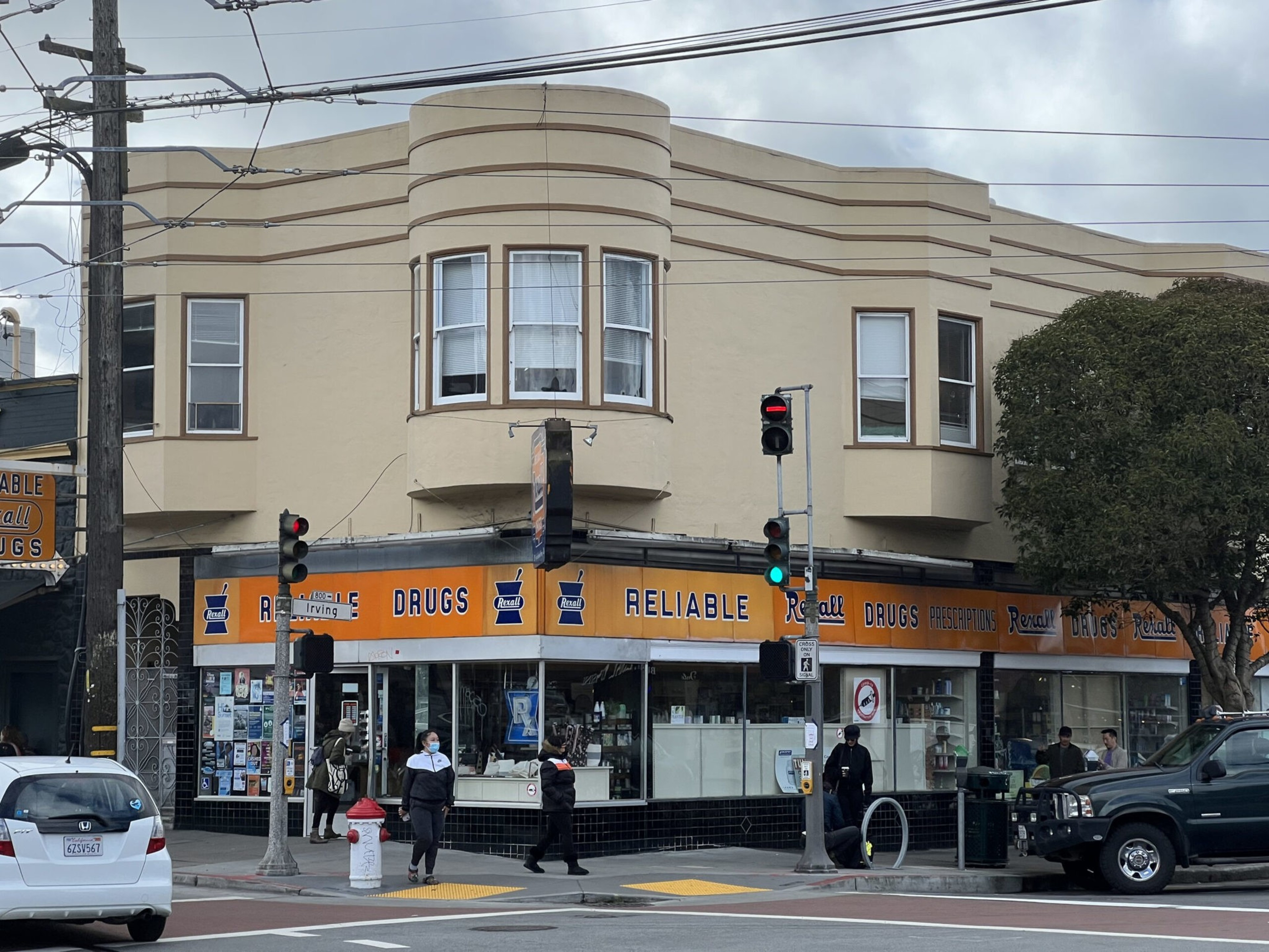 A beige corner building with a vintage orange Rexall Drugs sign wraps around the ground floor, with pedestrians crossing nearby and vehicles parked.