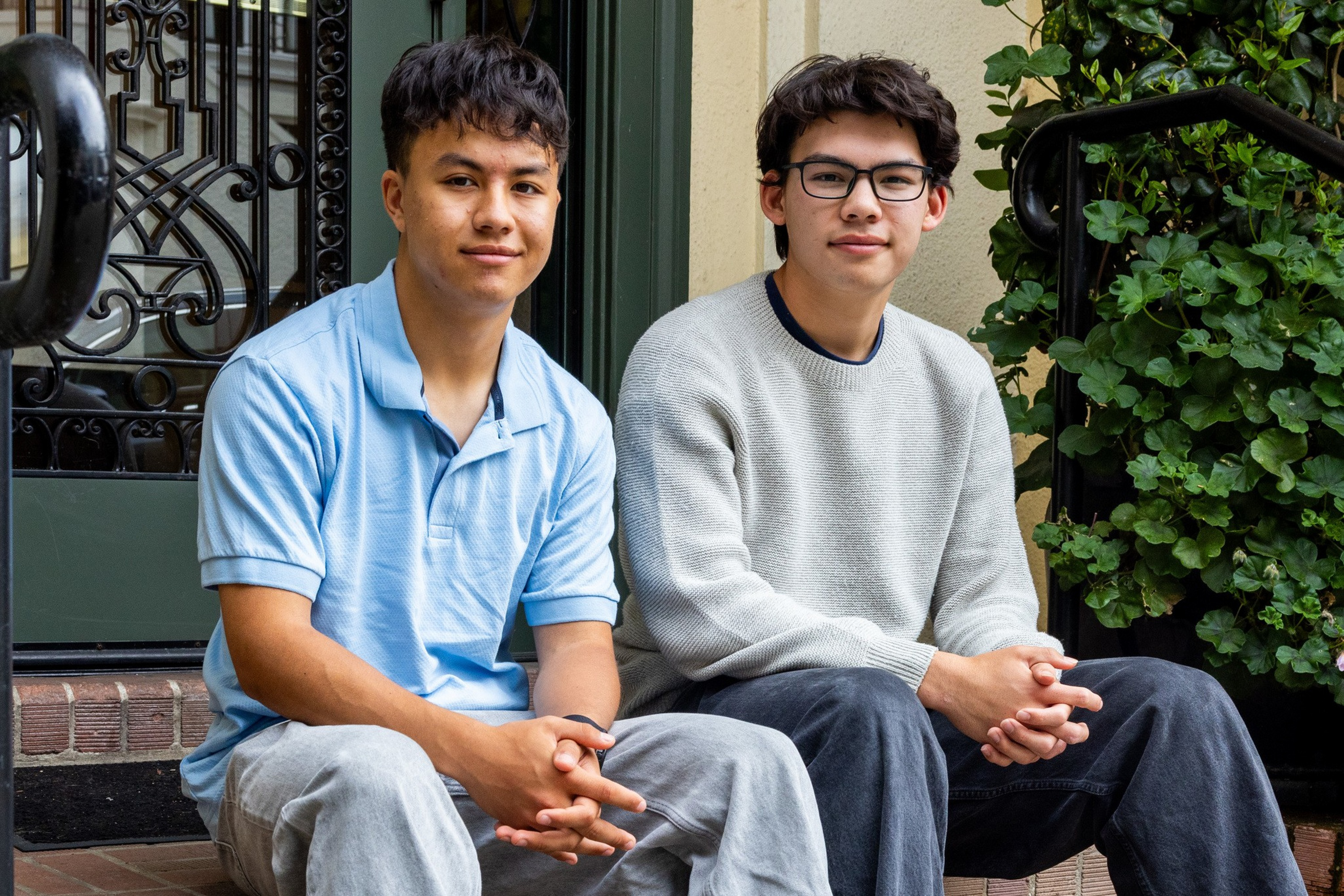 Two young men sit on steps in front of a door, one wearing a light blue polo and the other a gray sweater with glasses.
