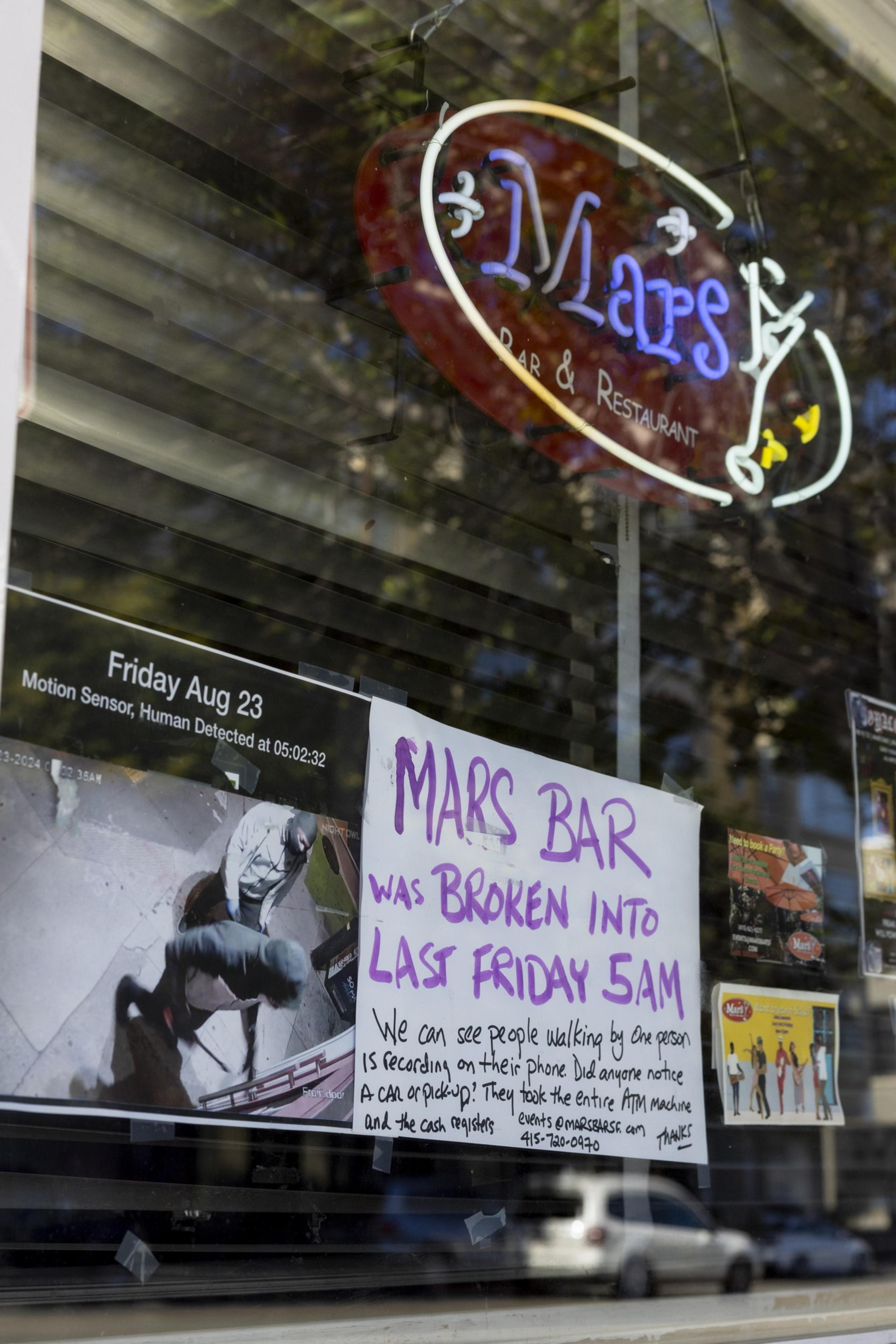 A neon &quot;Mars Bar &amp; Restaurant&quot; sign is seen over a surveillance image of a robbery. Below, a handwritten sign details the break-in, seeking further information from onlookers.