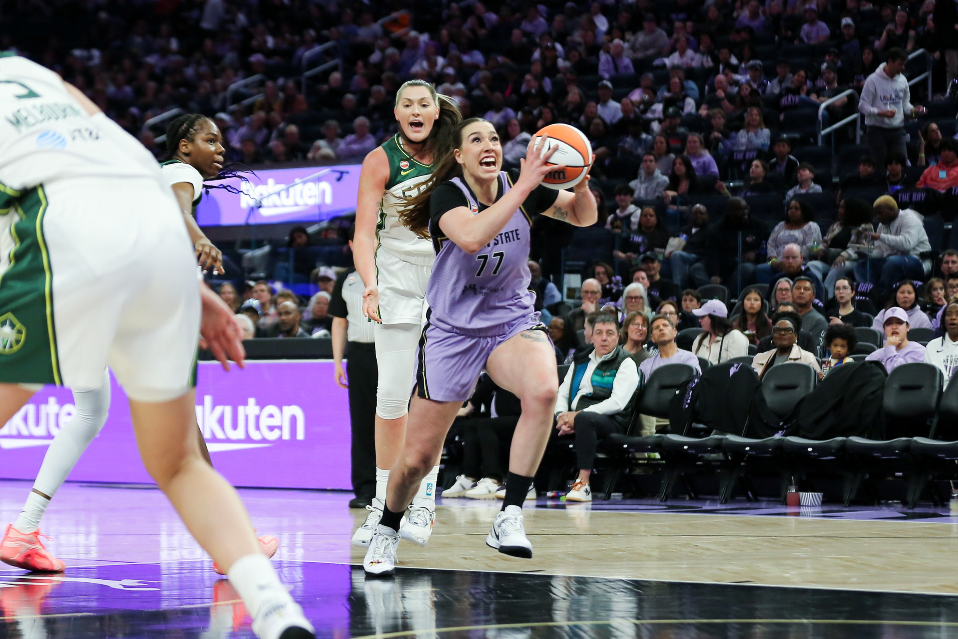A female basketball player in a purple jersey drives toward the basket with the ball, while defenders in white jerseys try to block her.