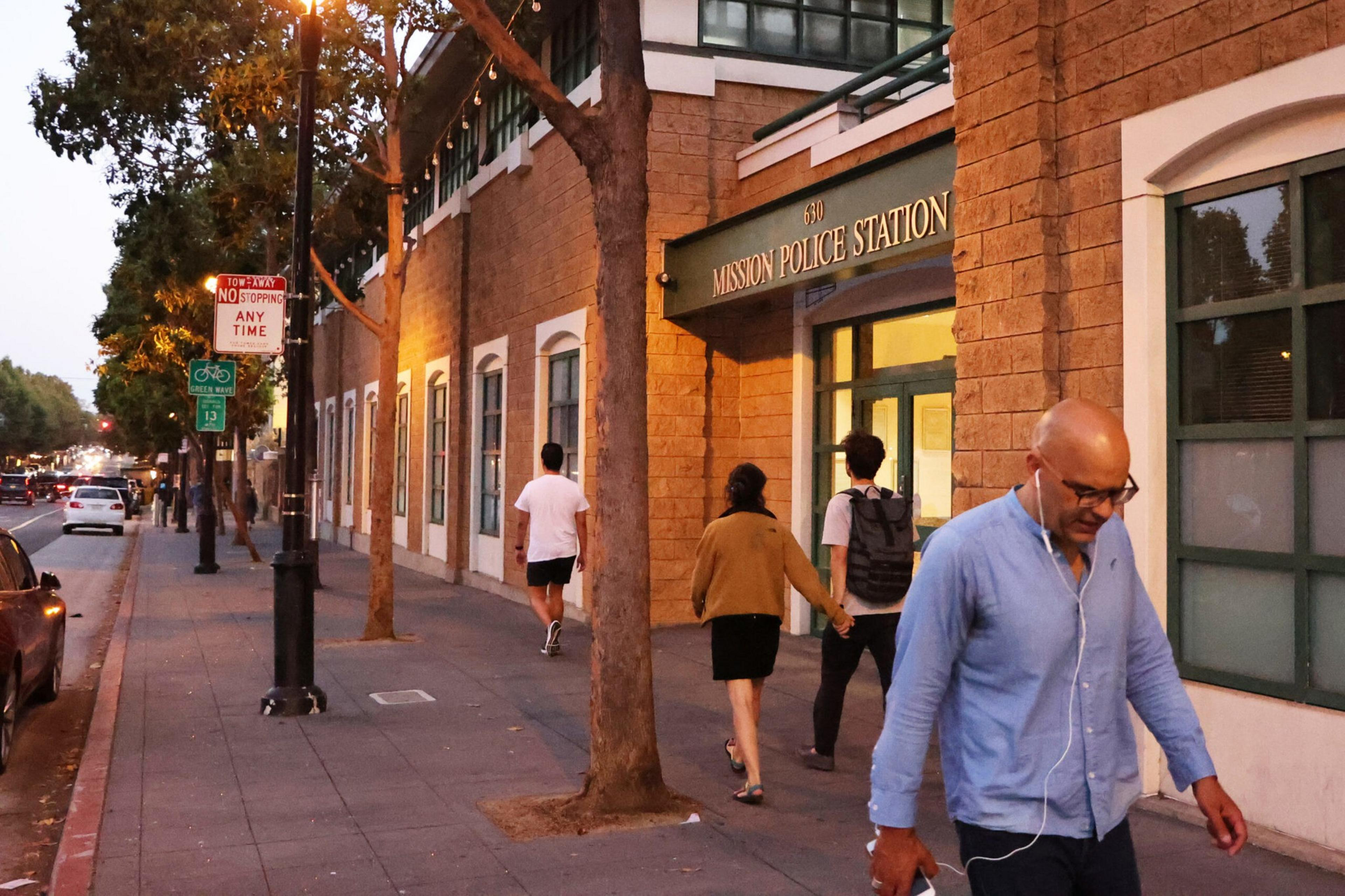 A man wearing earphones walks past the Mission Police Station as three people walk along the sidewalk during early evening.
