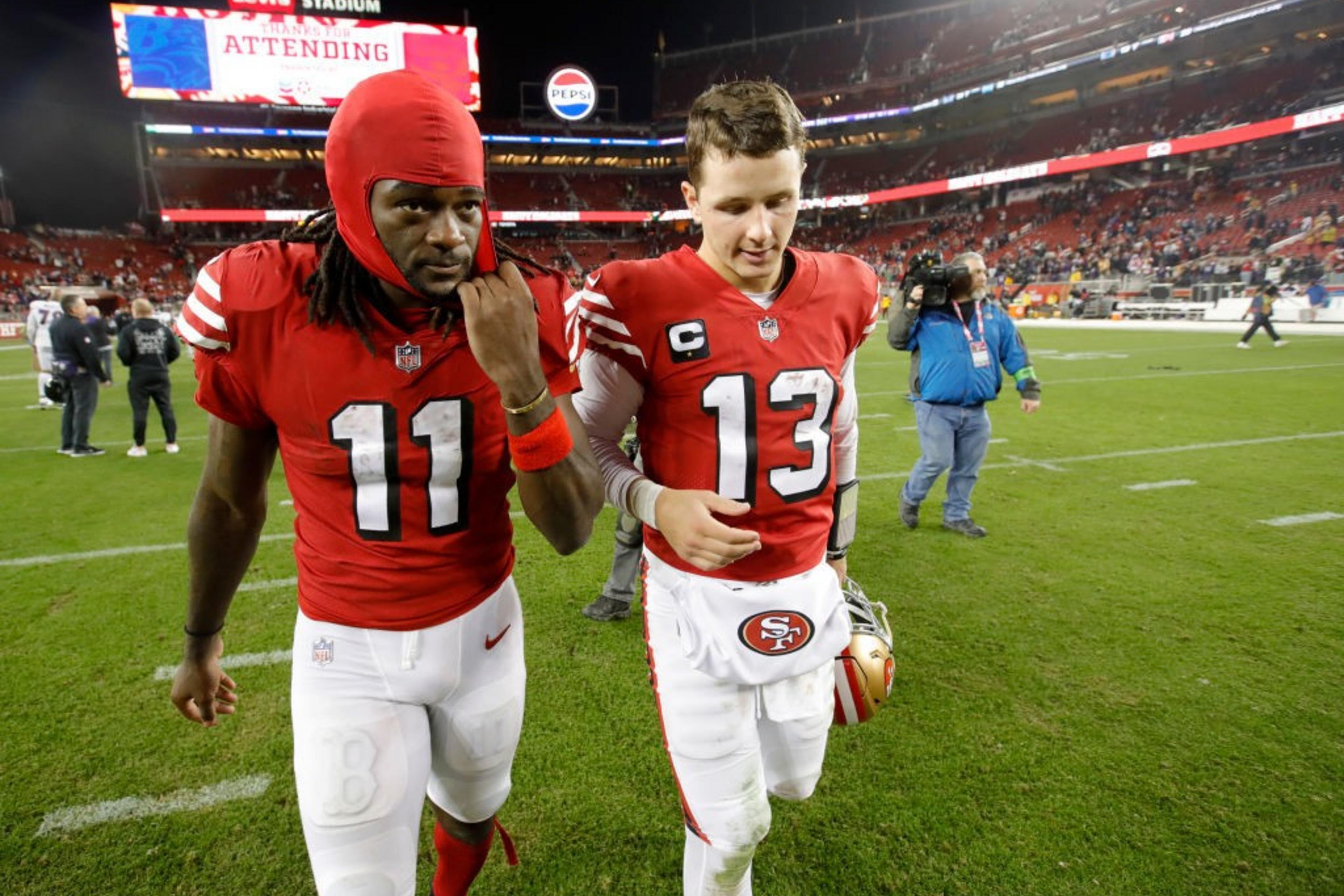 Two football players in red jerseys, numbered 11 and 13, walk together on a field. The stadium is bustling with people, and a large screen is in the background.