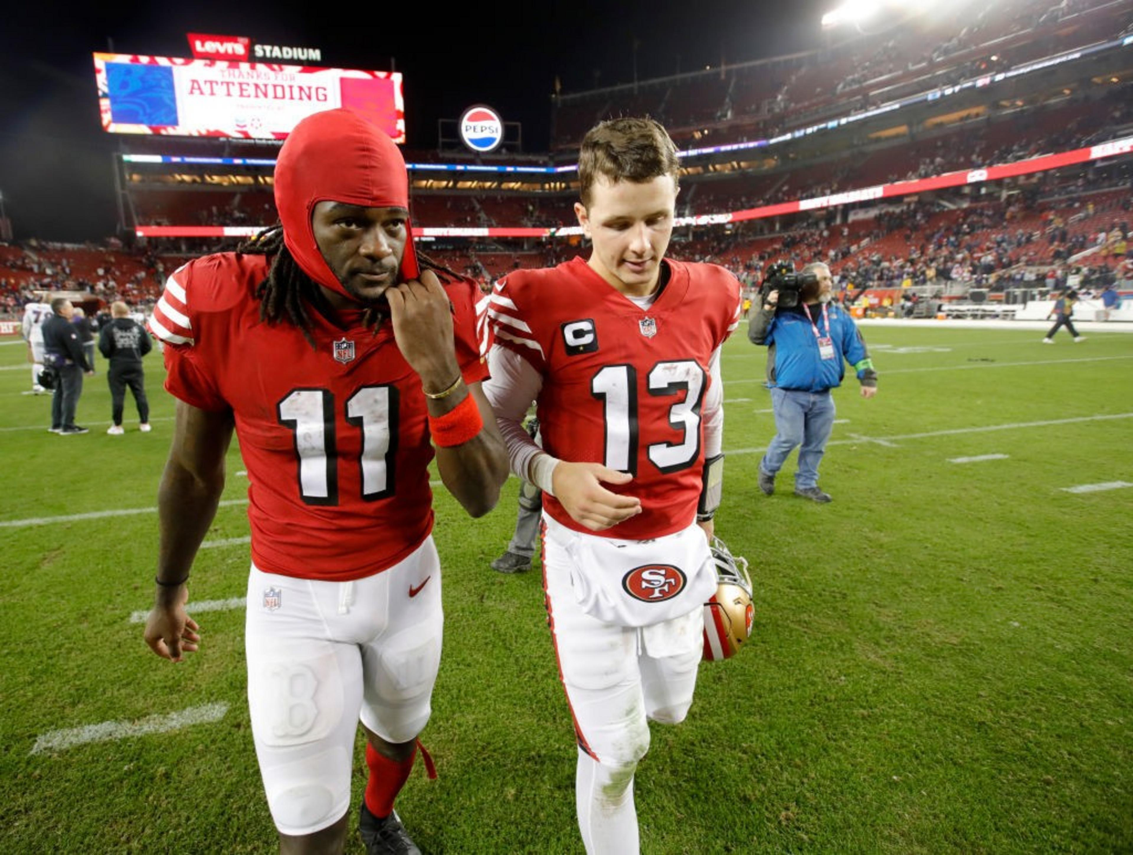 Two football players in red jerseys, numbered 11 and 13, walk together on a field. The stadium is bustling with people, and a large screen is in the background.