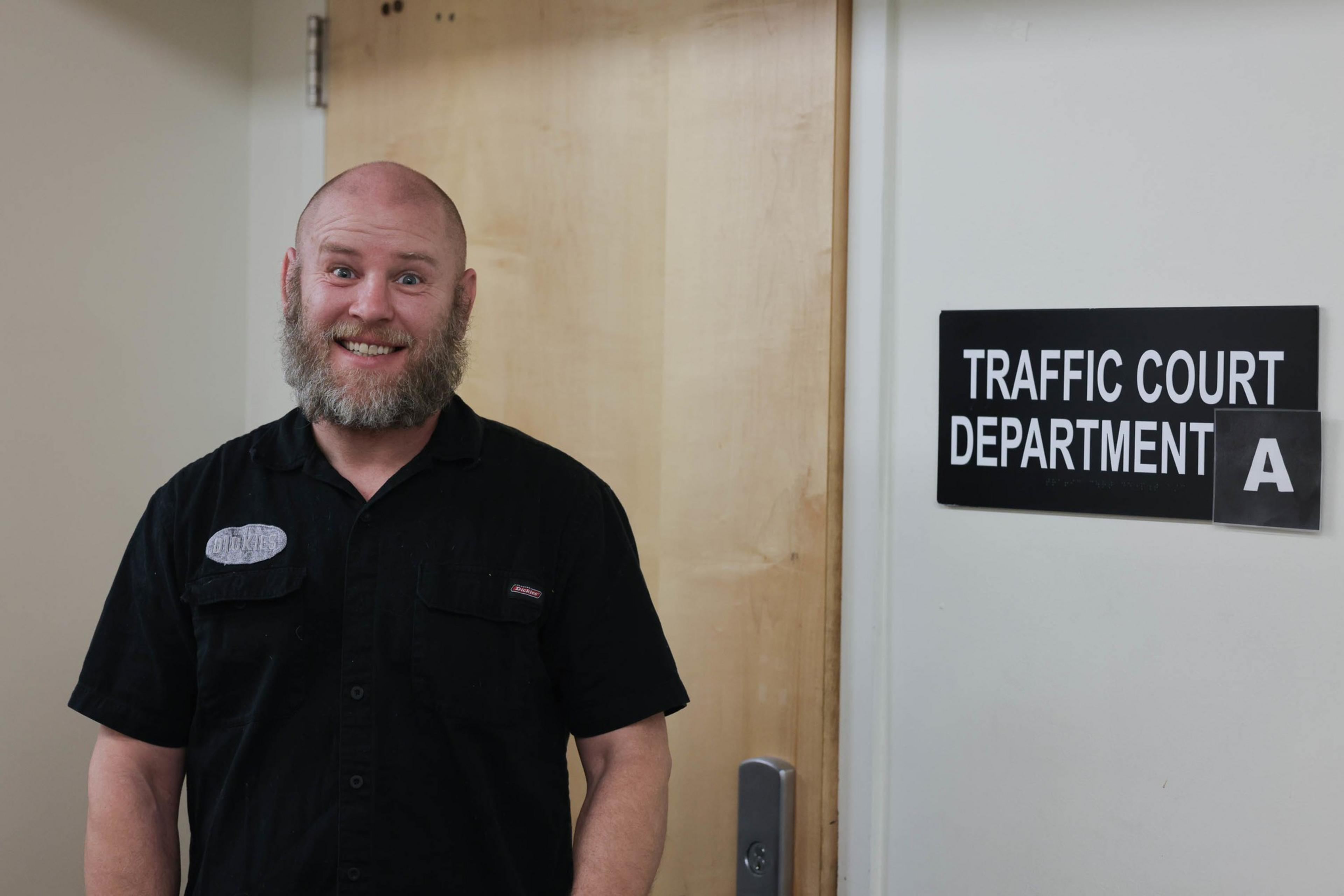 A person smiles next to a sign that reads &quot;traffic court department A&quot;.