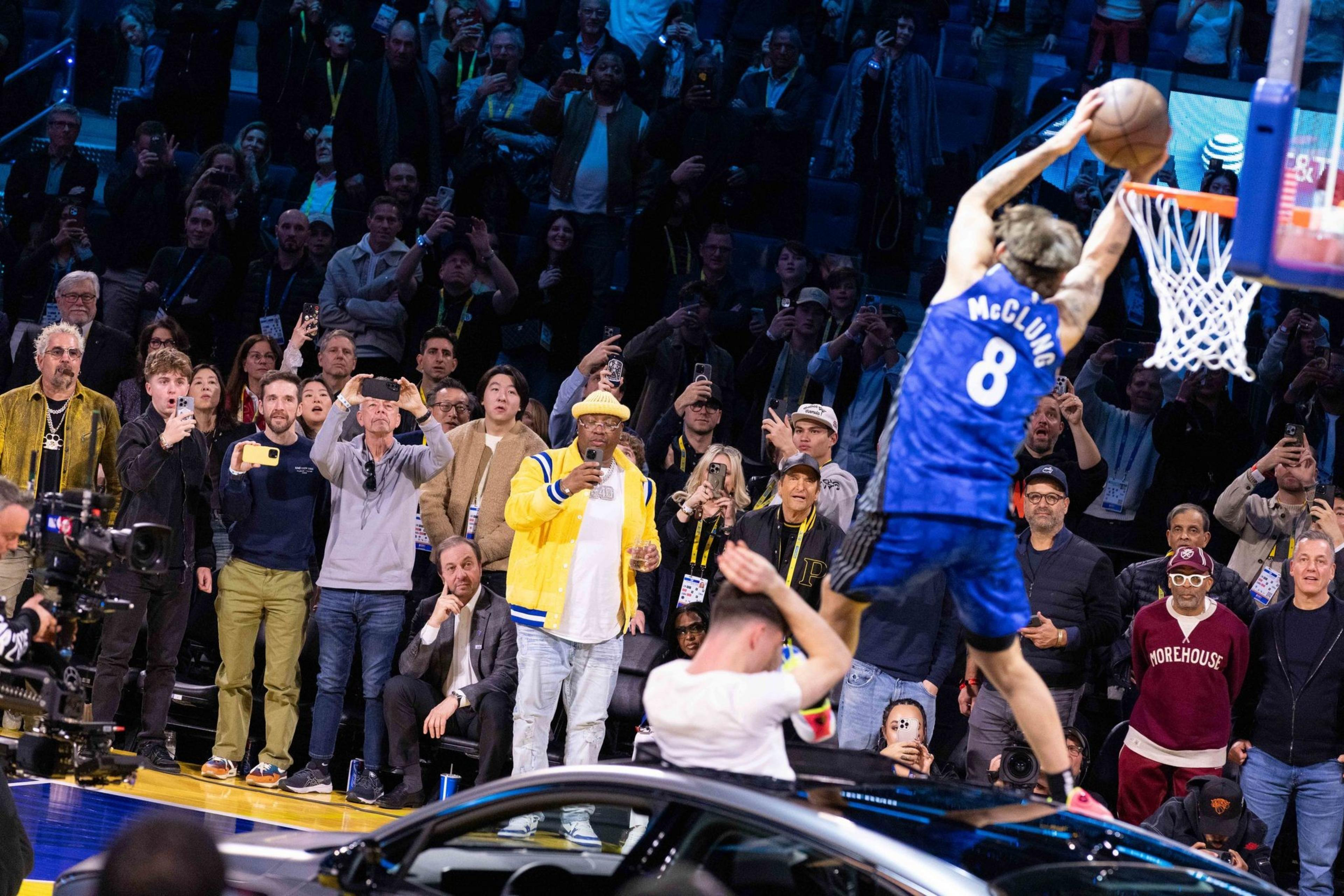 A basketball player wearing a blue jersey numbered 8 is mid-air dunking a ball into a hoop. A crowd is capturing the moment with phones, while a car is nearby.