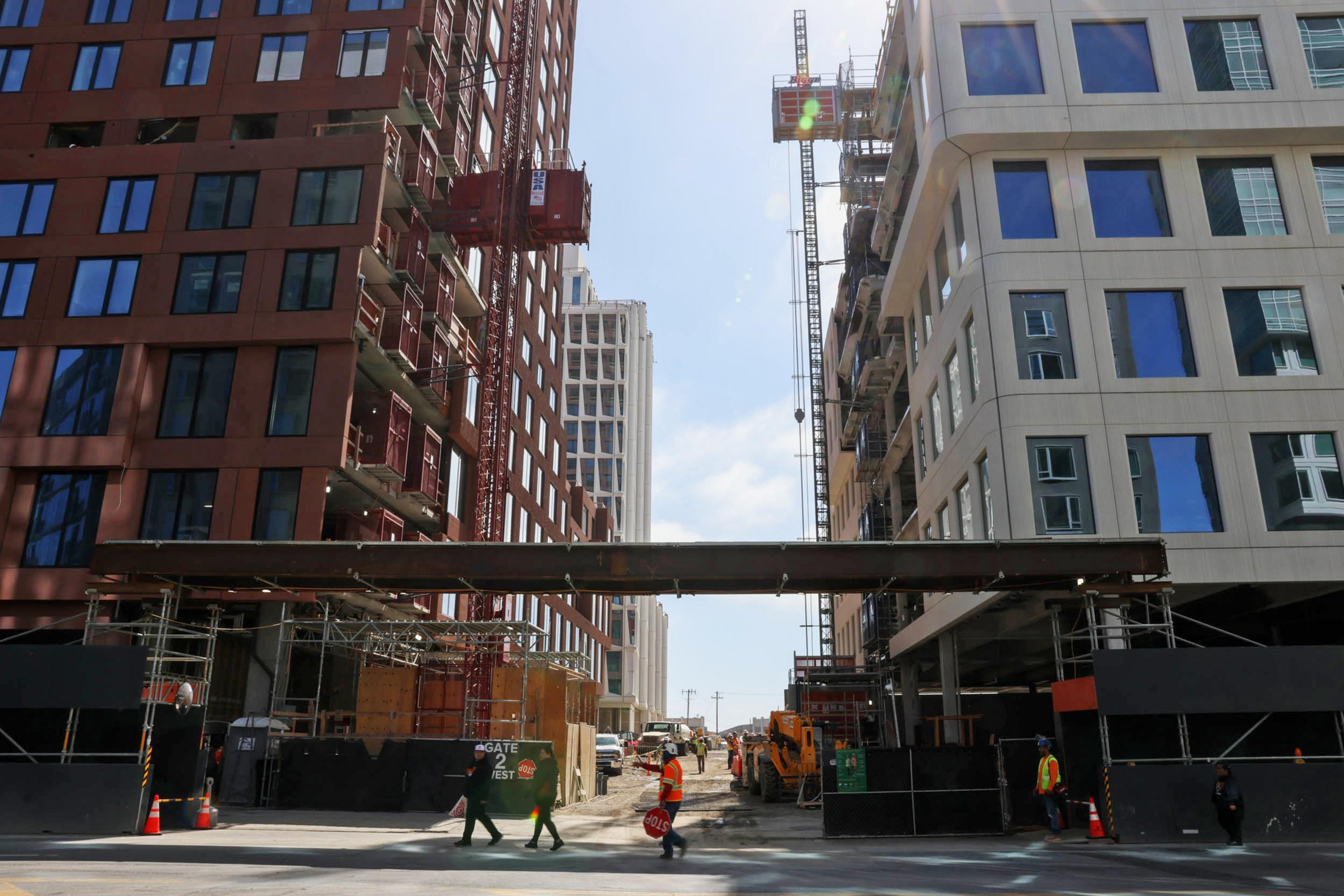Two tall buildings under construction flank a narrow street. Workers in safety gear are visible, and there's construction equipment and scaffolding present.