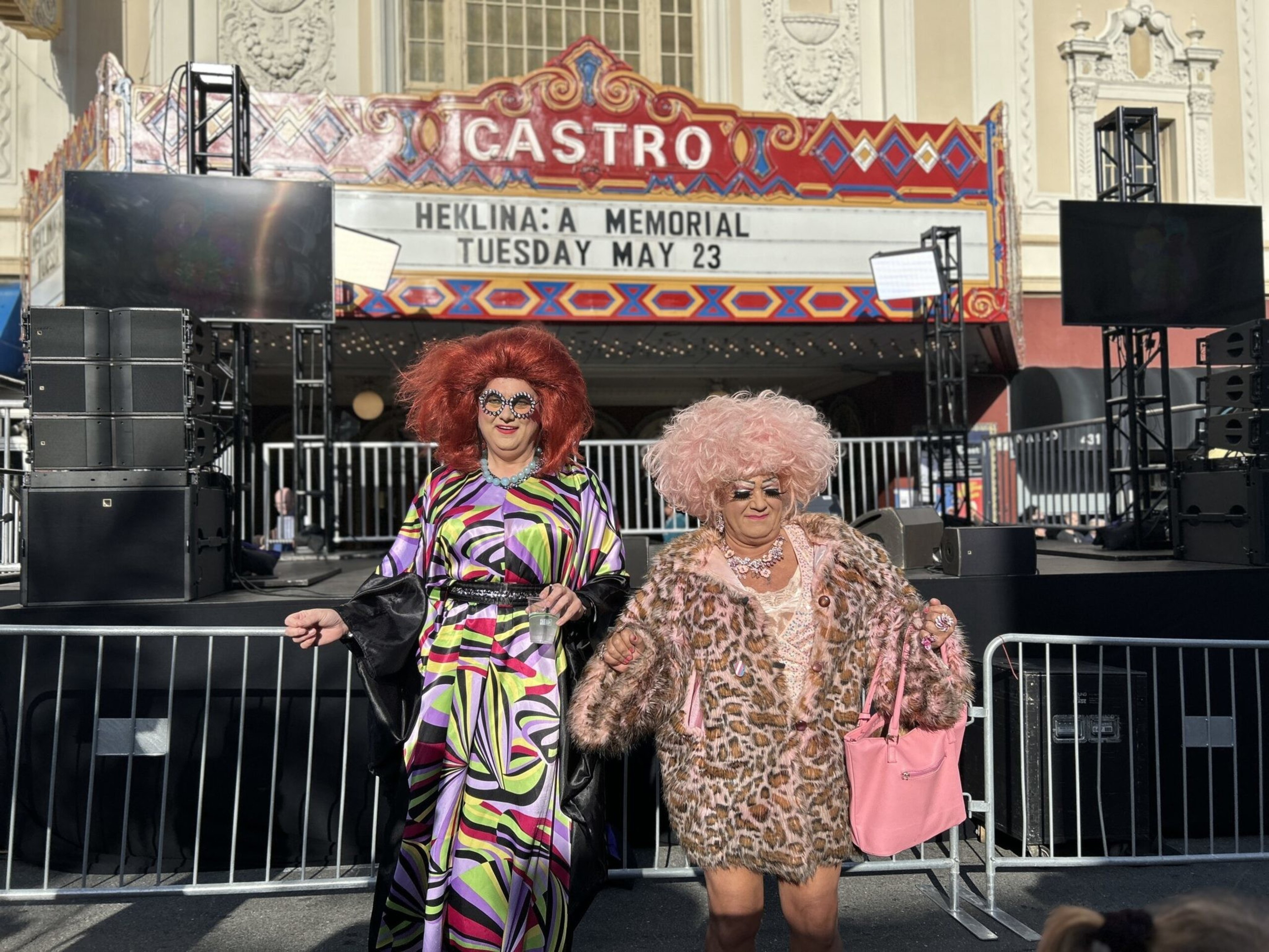 Two people in colorful, extravagant outfits pose happily in front of the Castro Theatre marquee announcing "Heklina: A Memorial" on May 23.