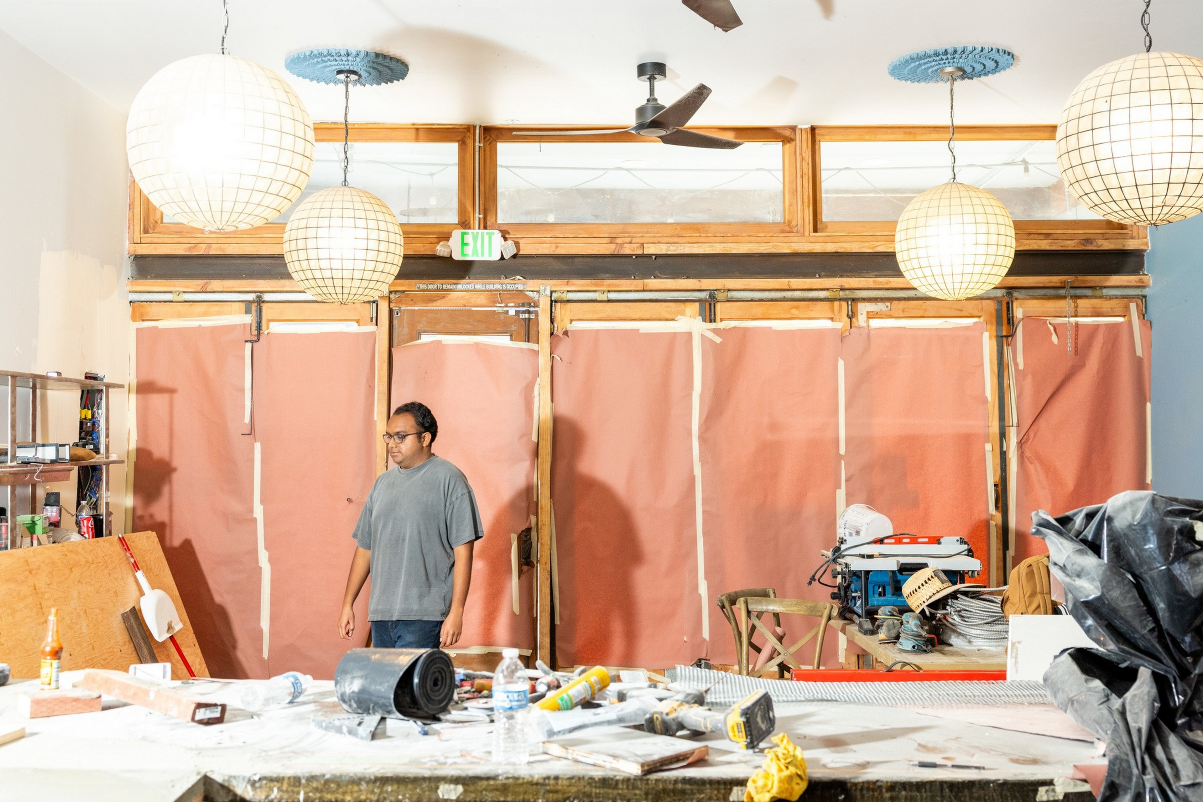 A person stands in a cluttered workshop with hanging paper lanterns, tools, and materials scattered on tables, and pinkish sheets taped on the back wall.