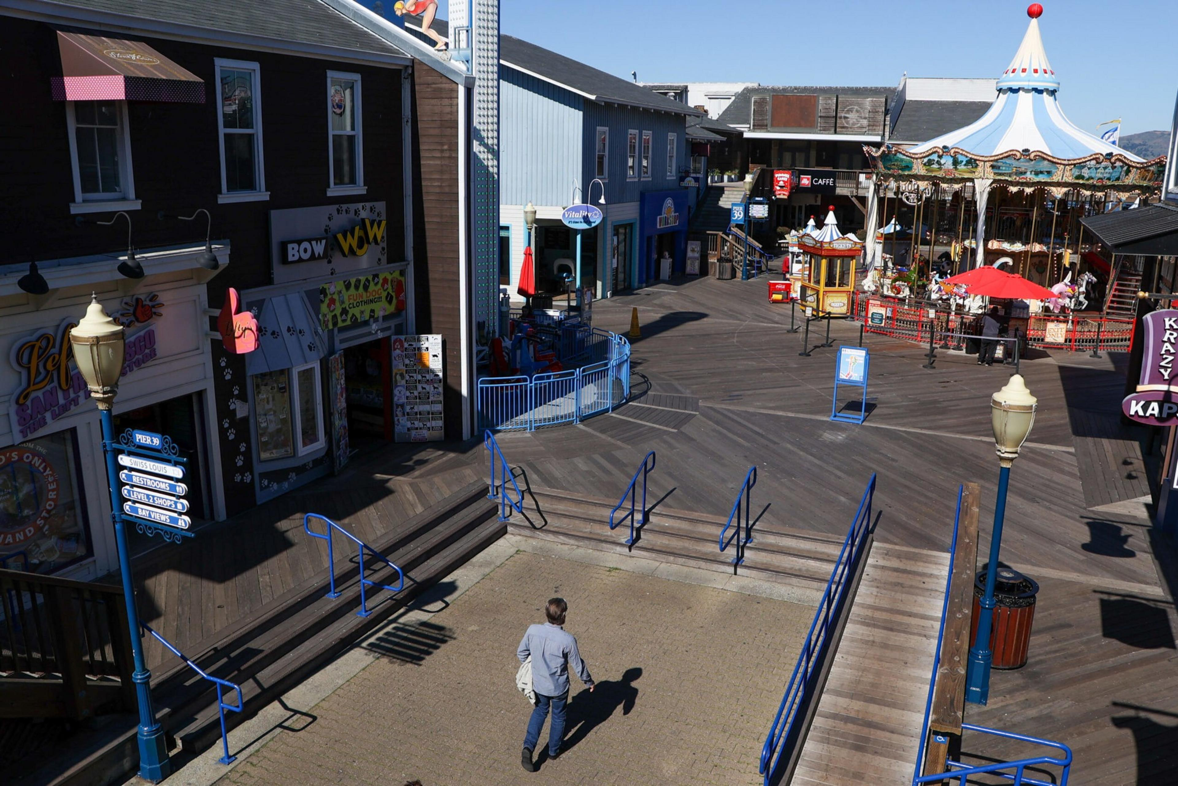 A person walks alone on a wooden boardwalk surrounded by colorful shops and a carousel under a clear blue sky.
