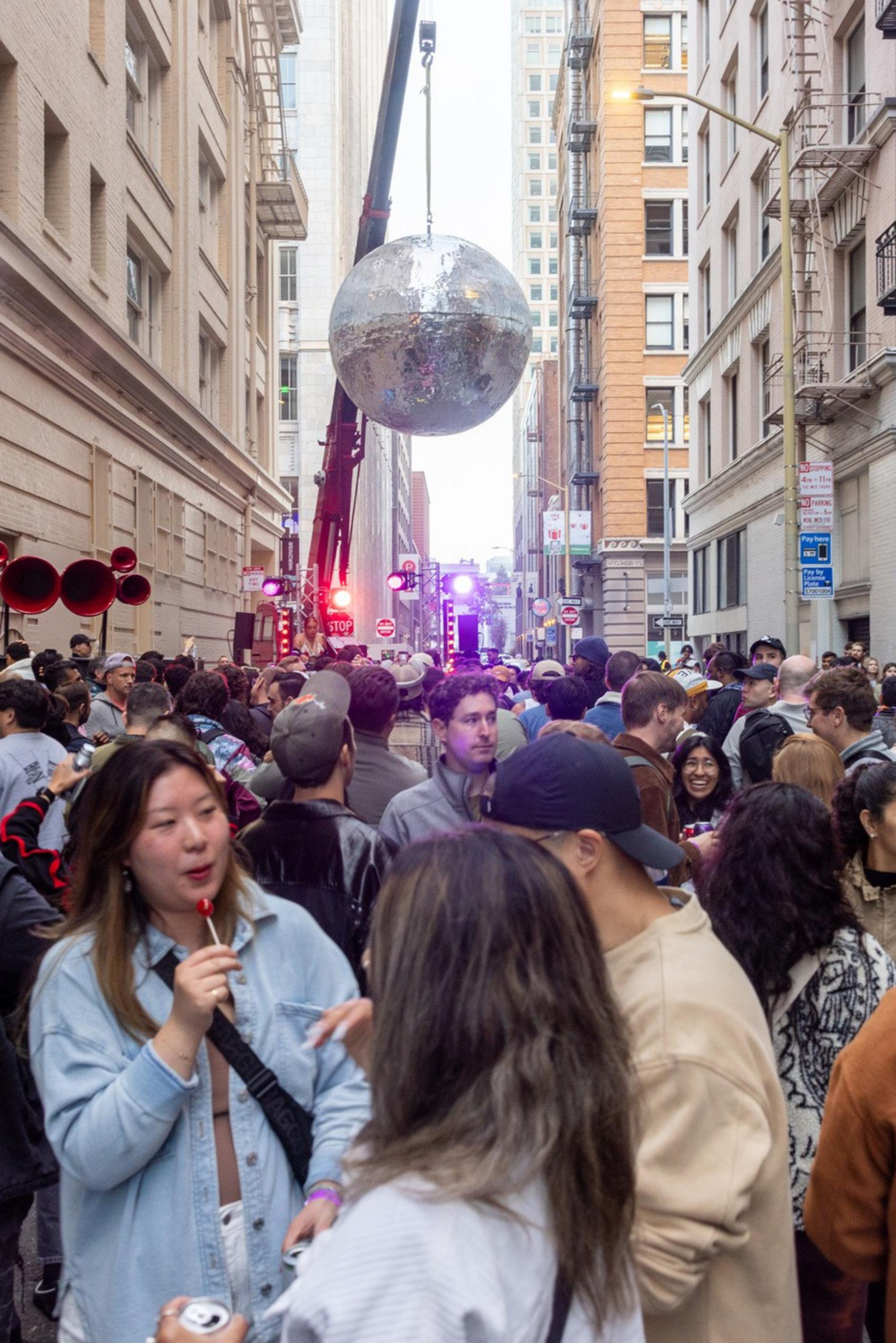 A crowded street party in a city with people mingling, eating, and talking. Large silver disco ball hanging mid-street. Buildings line both sides, creating an alley-like effect.