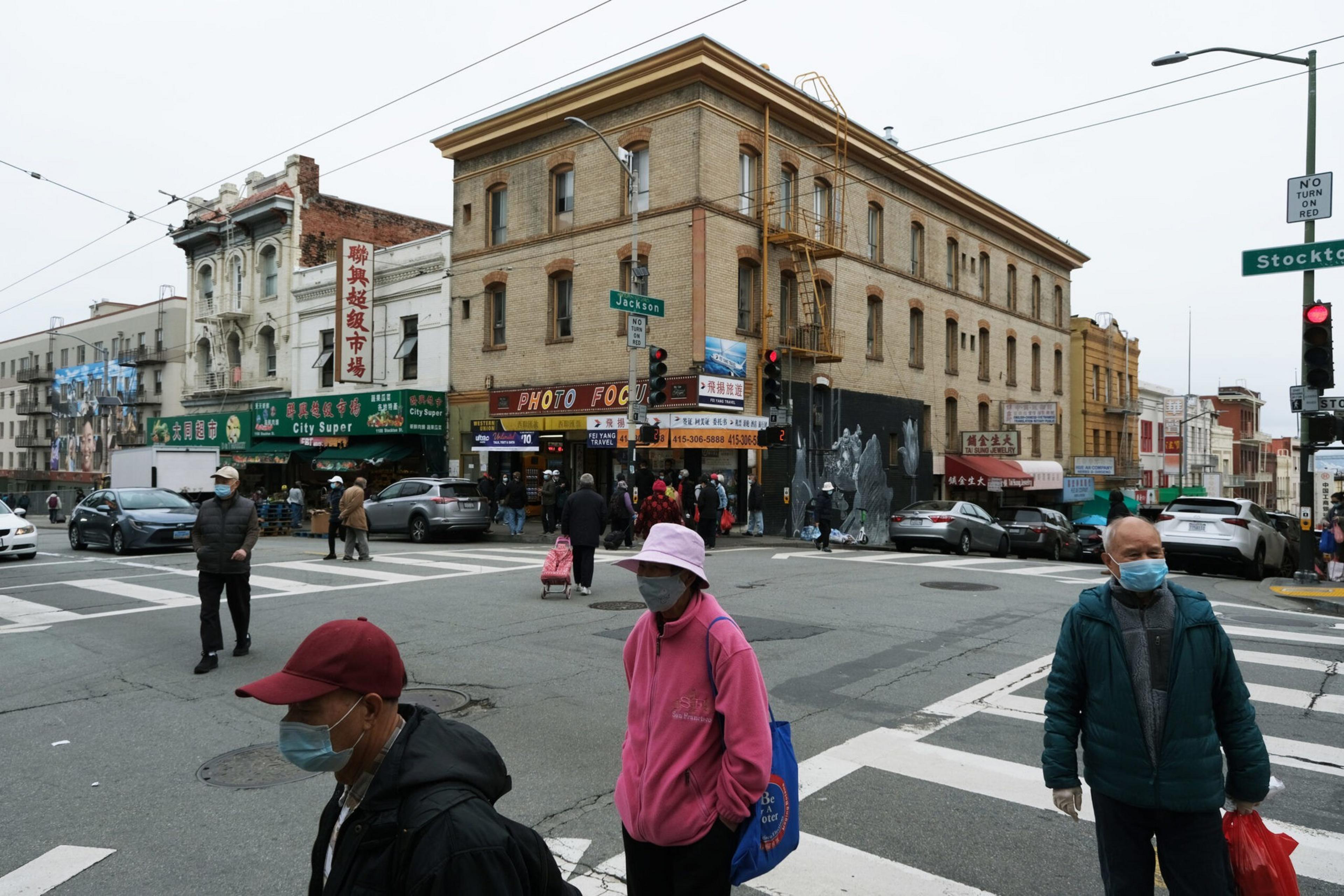 People wearing face masks walk on a city street corner with older buildings, stores including a supermarket and a photo shop with signs in English and Chinese.