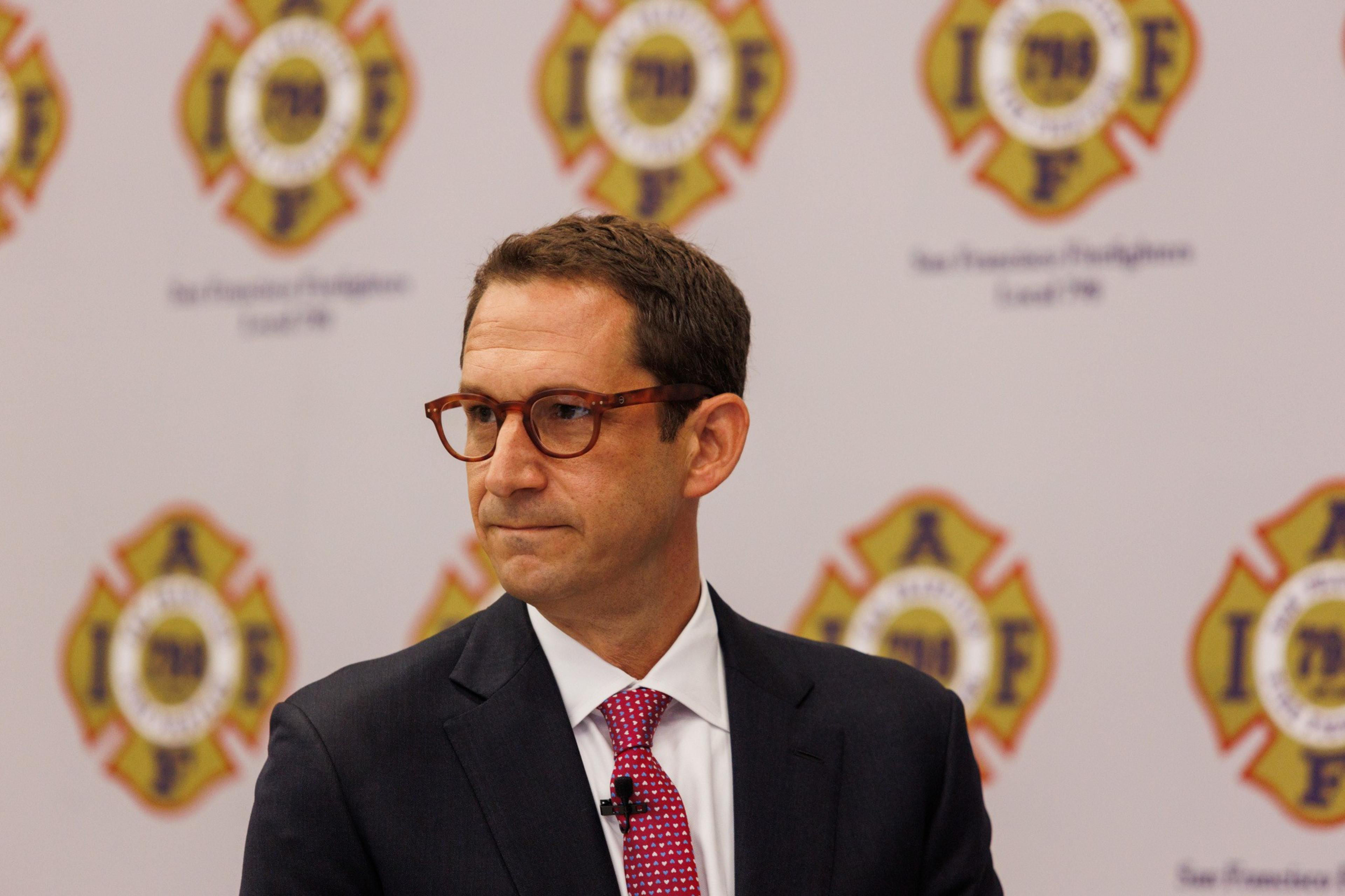 A man in a suit and red tie stands in front of a backdrop with fire department logos, looking to the side. The logos read "San Francisco Firefighters Local 798."