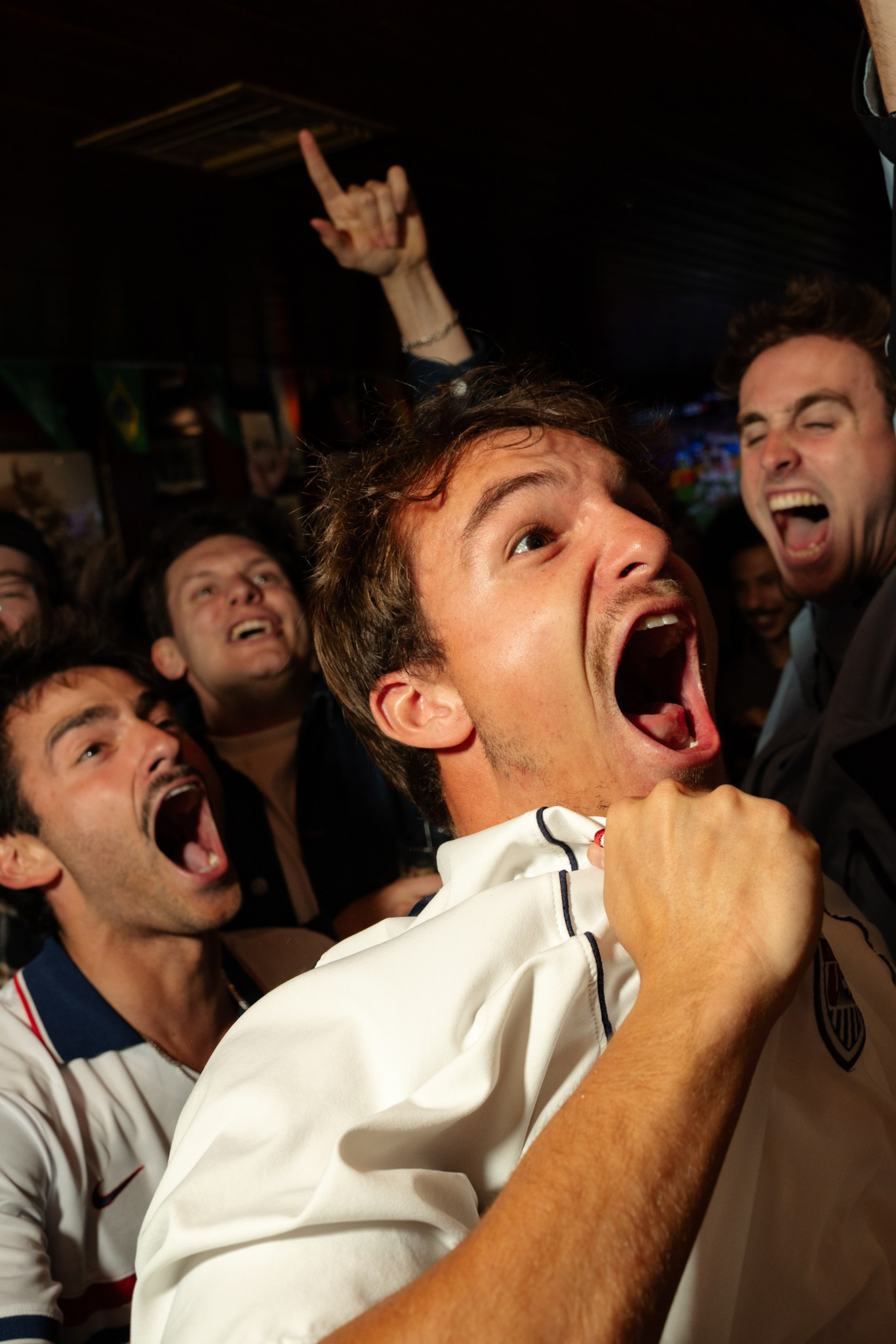 Several young men are shouting and cheering loudly, with one in the foreground gripping his shirt in excitement, likely celebrating a sports event.