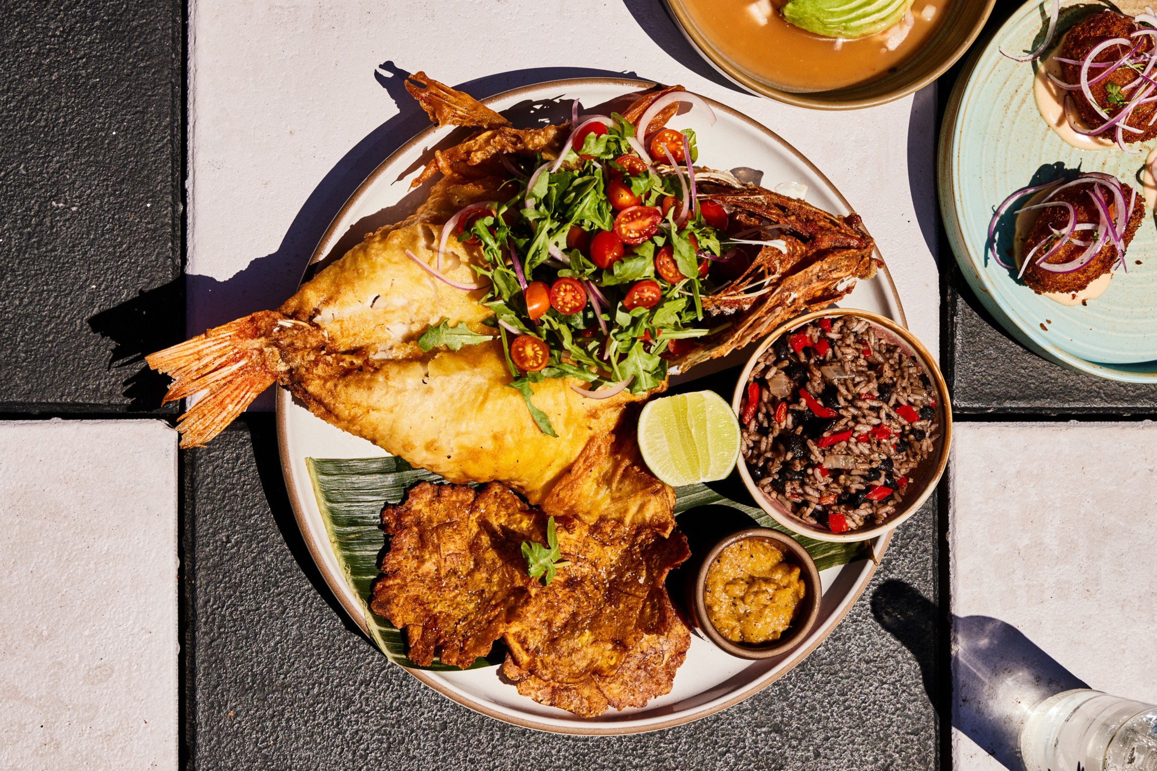 The image shows a plate with fried fish topped with a fresh salad of greens and cherry tomatoes, served with rice and beans, a lime wedge, and a small bowl of sauce.