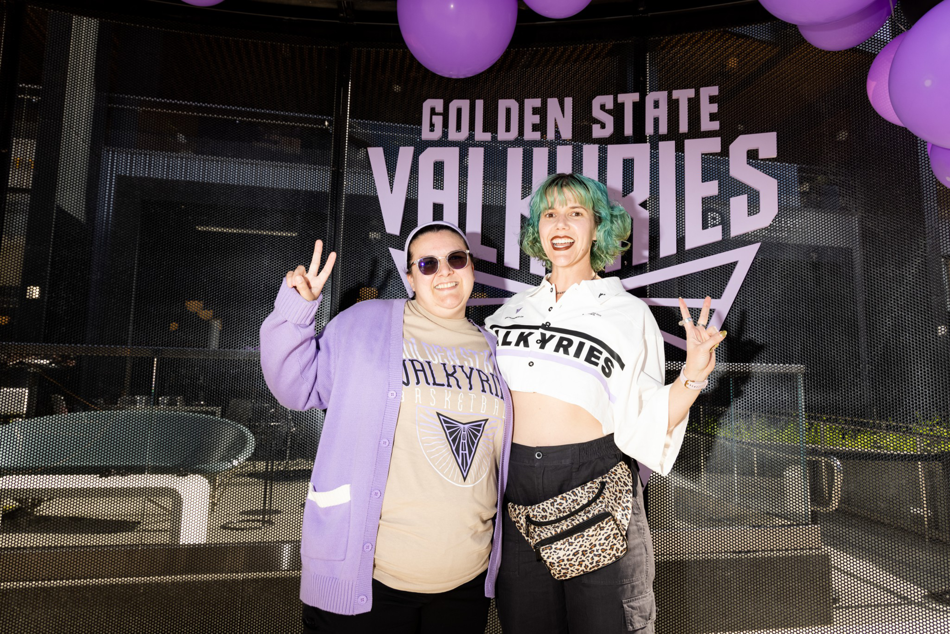 Two smiling people pose with peace signs under purple balloons in front of a “Golden State Valkyries” sign, wearing matching team-themed clothing.