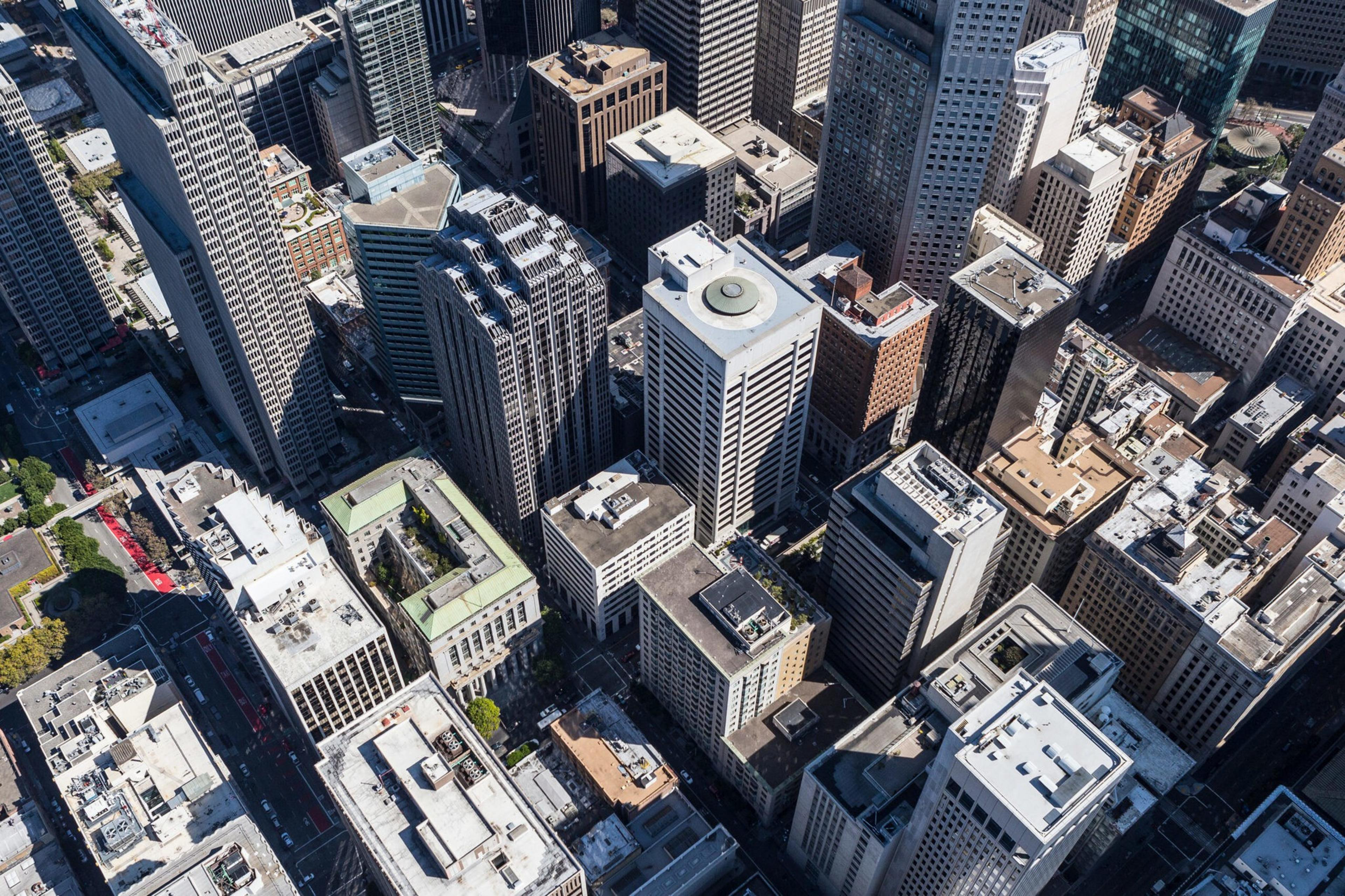 A dense cluster of skyscrapers and tall buildings casts shadows over a city street grid on a sunny day.