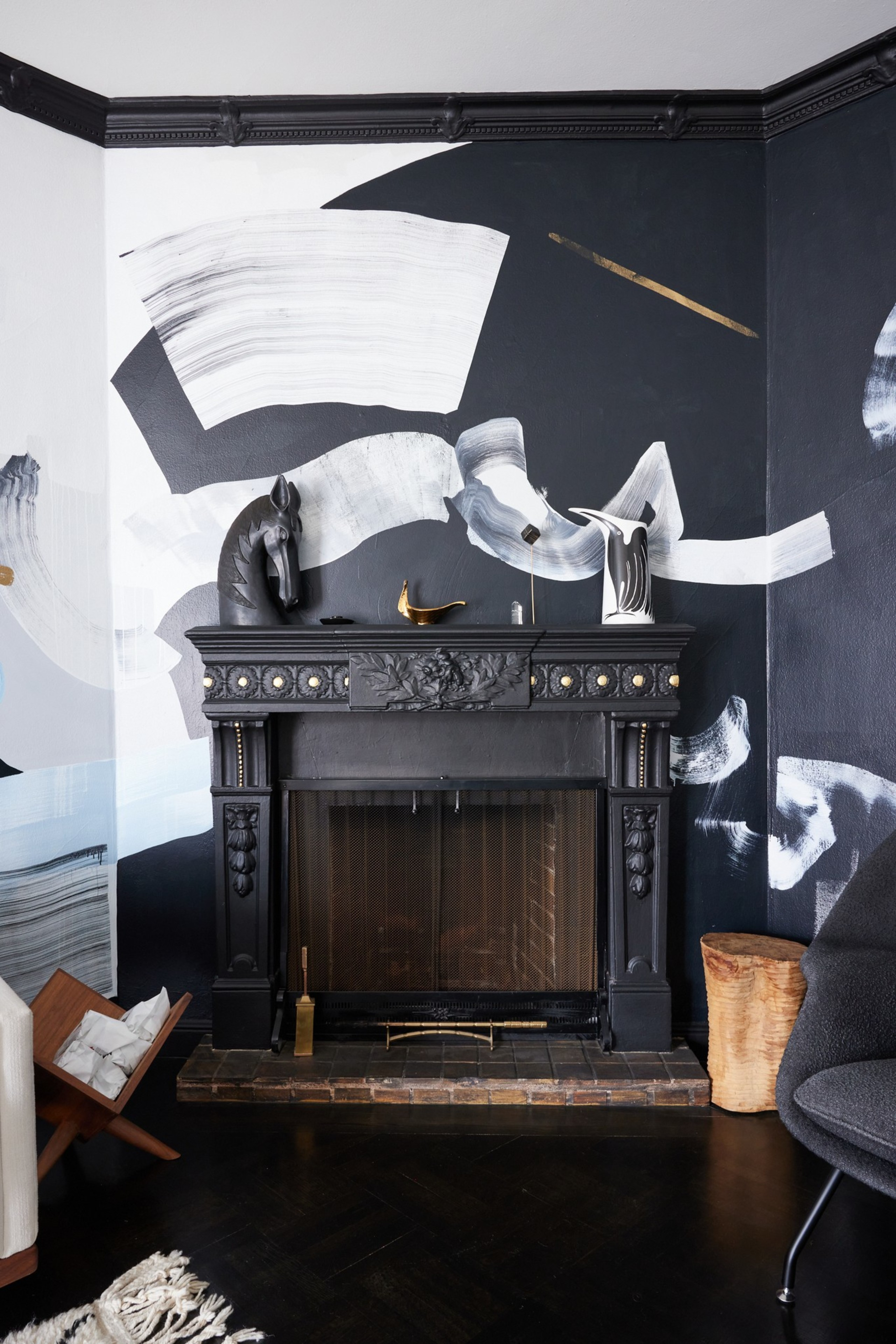 The image shows a stylish living room with a black ornate fireplace, artistic wall designs in black and white, modern decor pieces on the mantel, and dark wooden flooring.