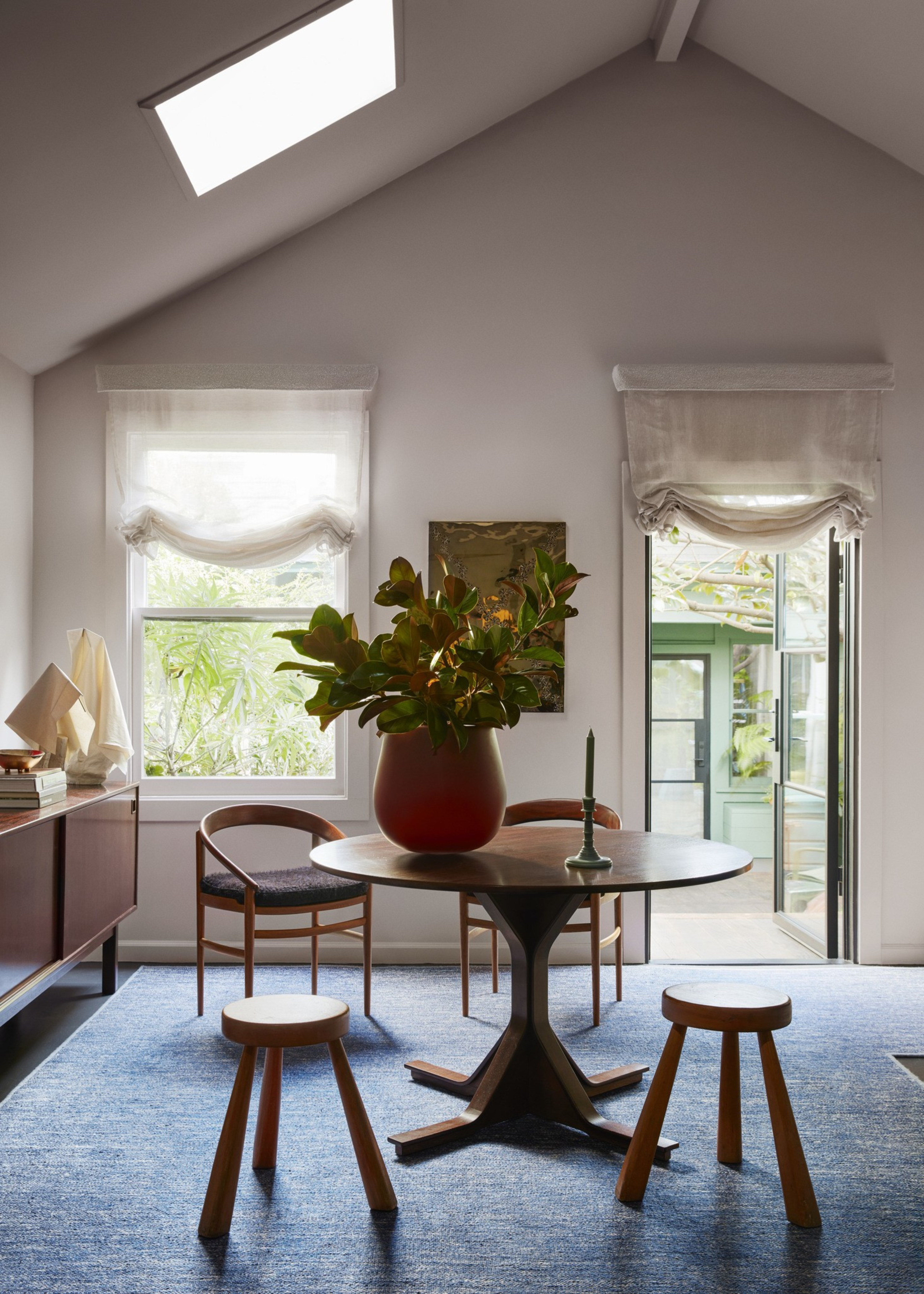 A round wooden table with a red vase holding green leaves is centered on a blue carpet, surrounded by two stools and two chairs near tall windows and a glass door.