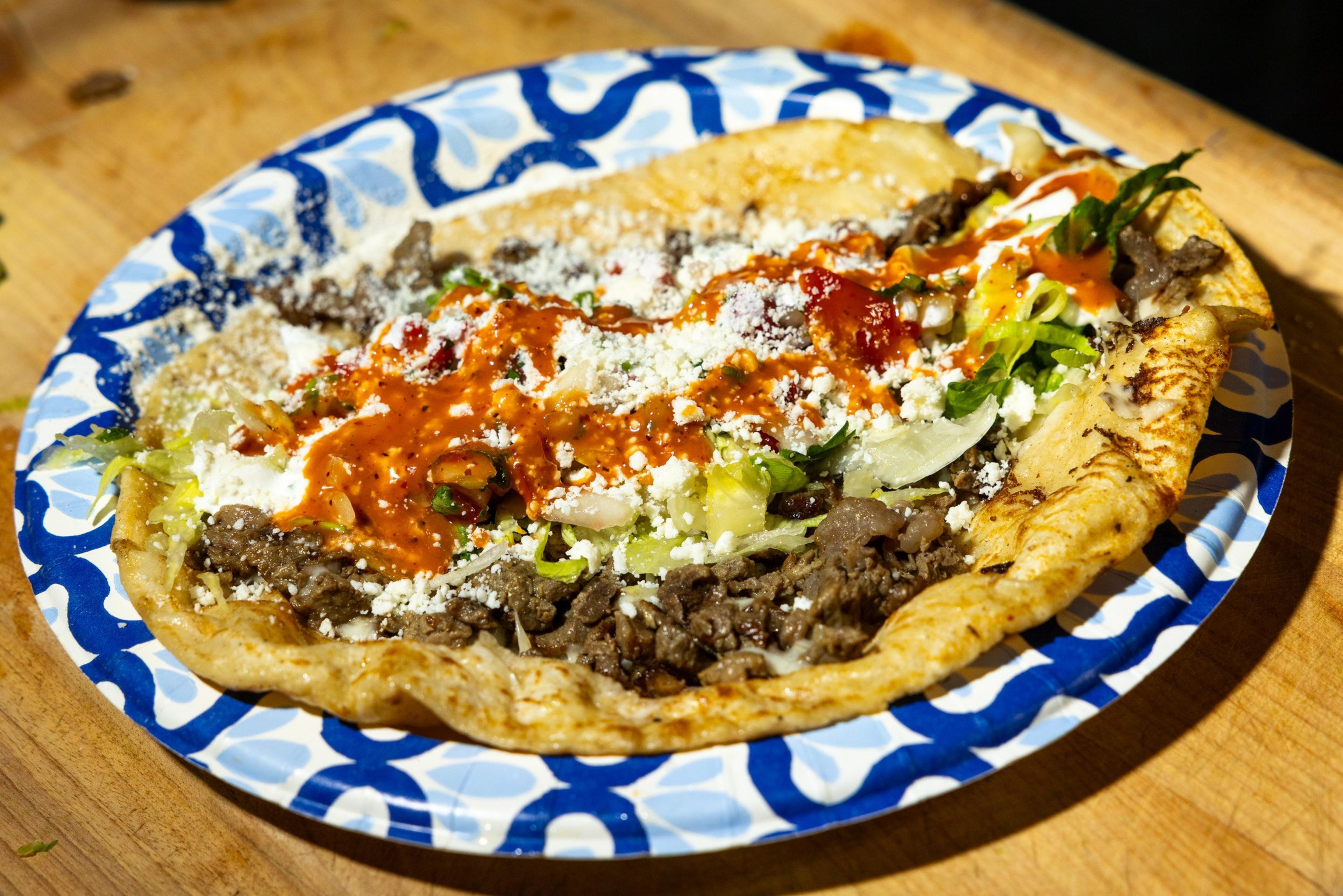 The image shows a taco or tostada on a decorative blue and white plate. It has toppings including beef, shredded lettuce, white cheese, salsa, and sour cream.