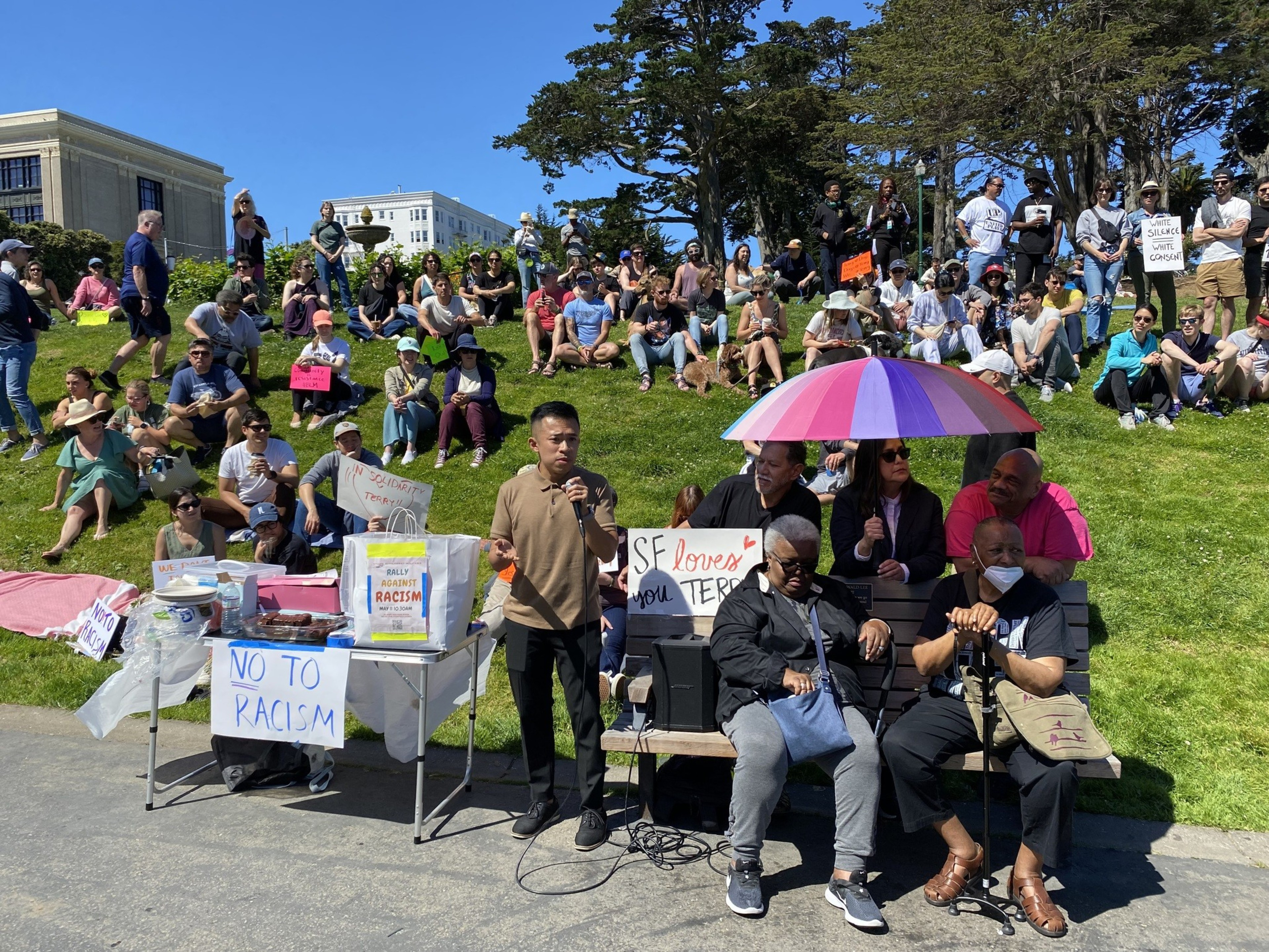 A diverse group of people sits on grass at an outdoor rally against racism with signs and a speaker.