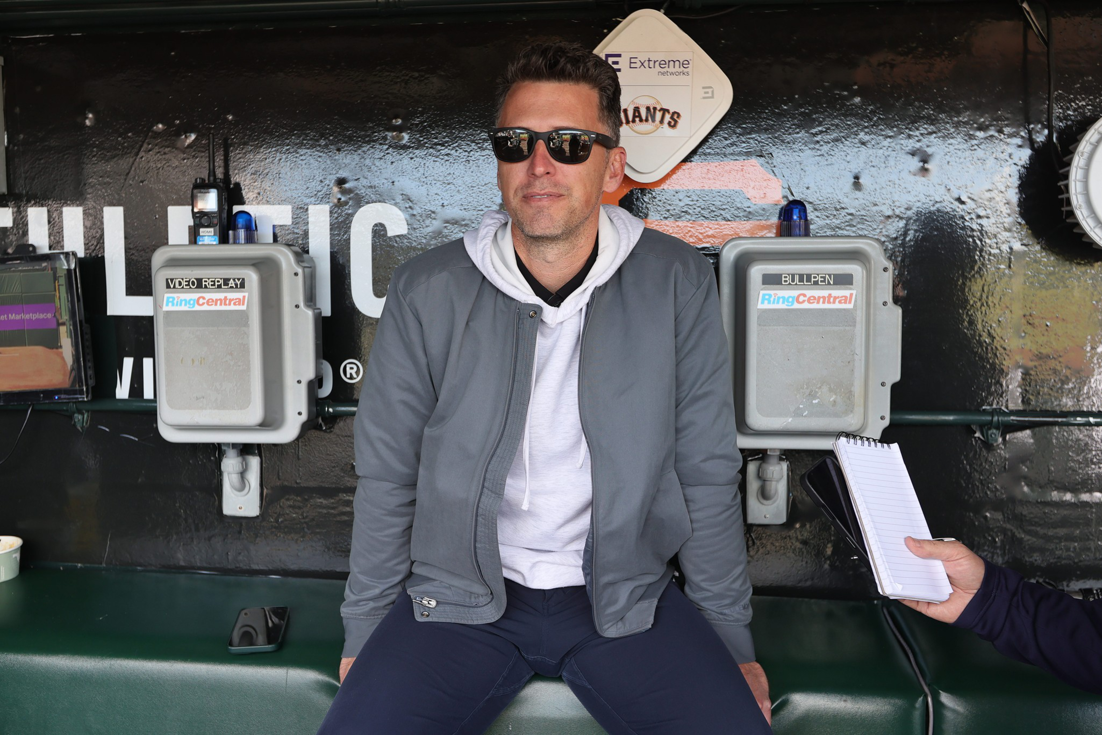 A man in sunglasses, a gray jacket, and white hoodie sits on a bench between two call boxes labeled “Video Replay” and “Bullpen.” A hand holds a notepad.