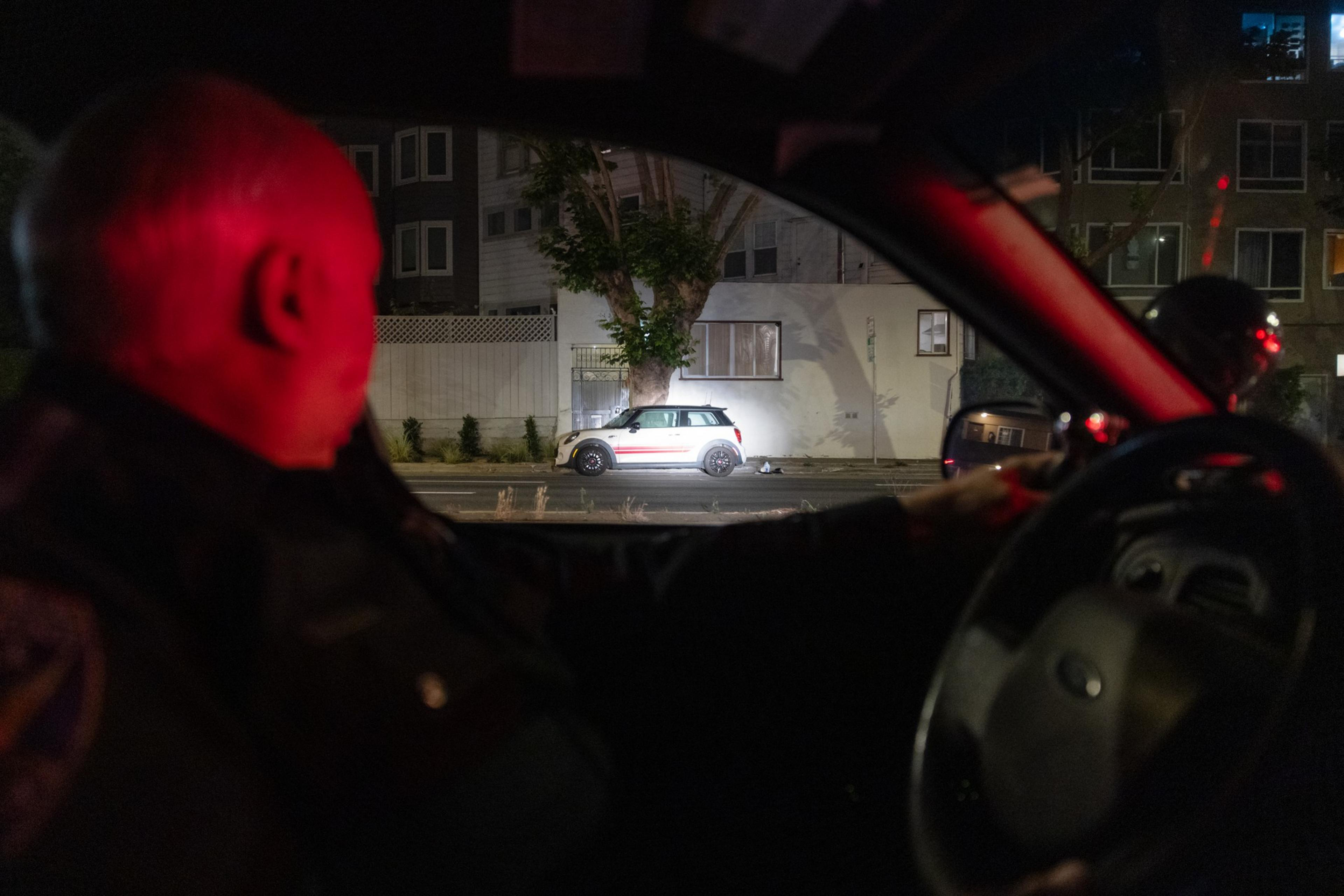 A police officer sits in a patrol car at night, looking out the window at a lit white Mini Cooper parked on the street.
