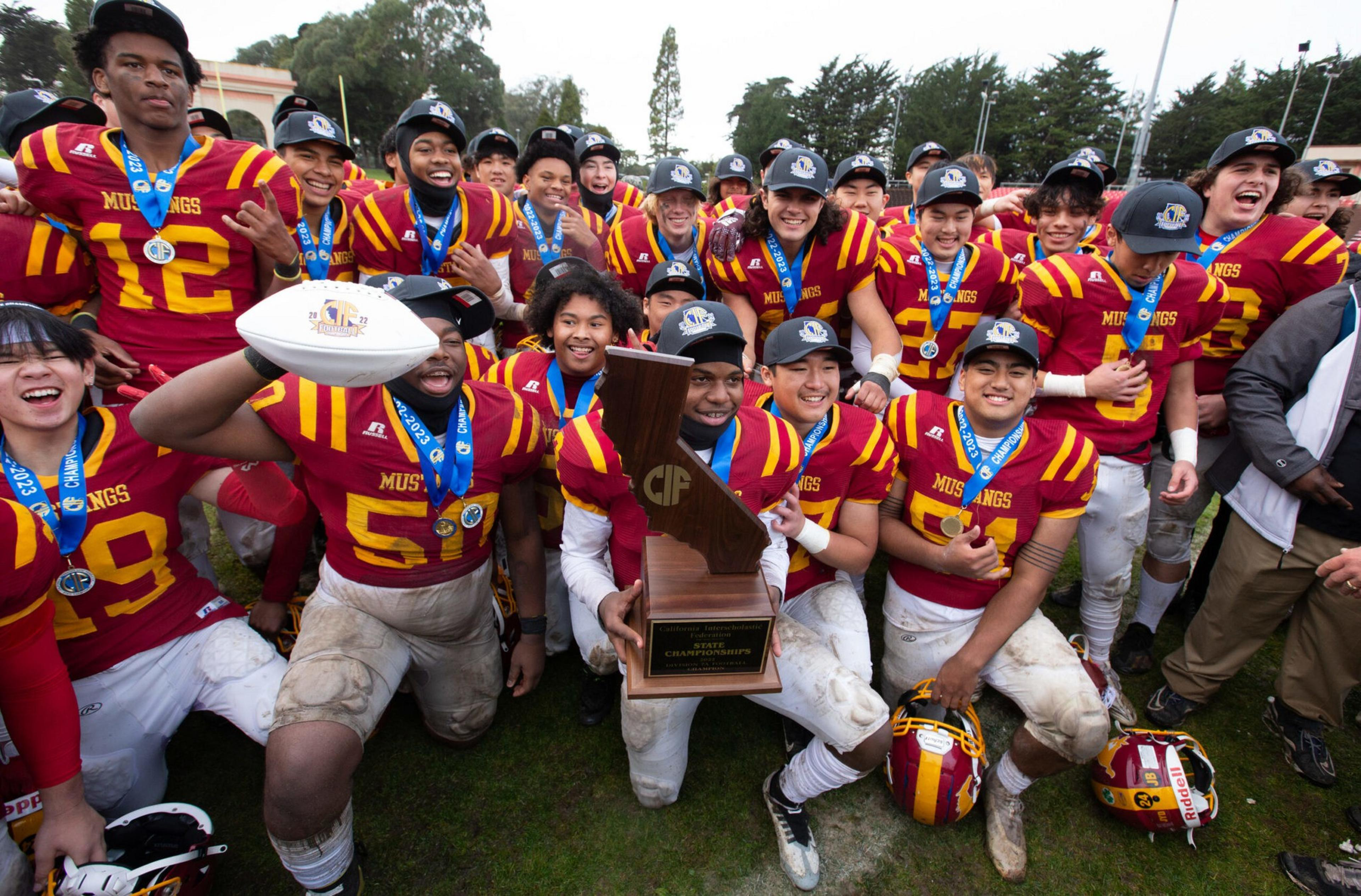 A football team in red and yellow uniforms celebrates, wearing medals and holding a large wooden trophy and a football on a grassy field.