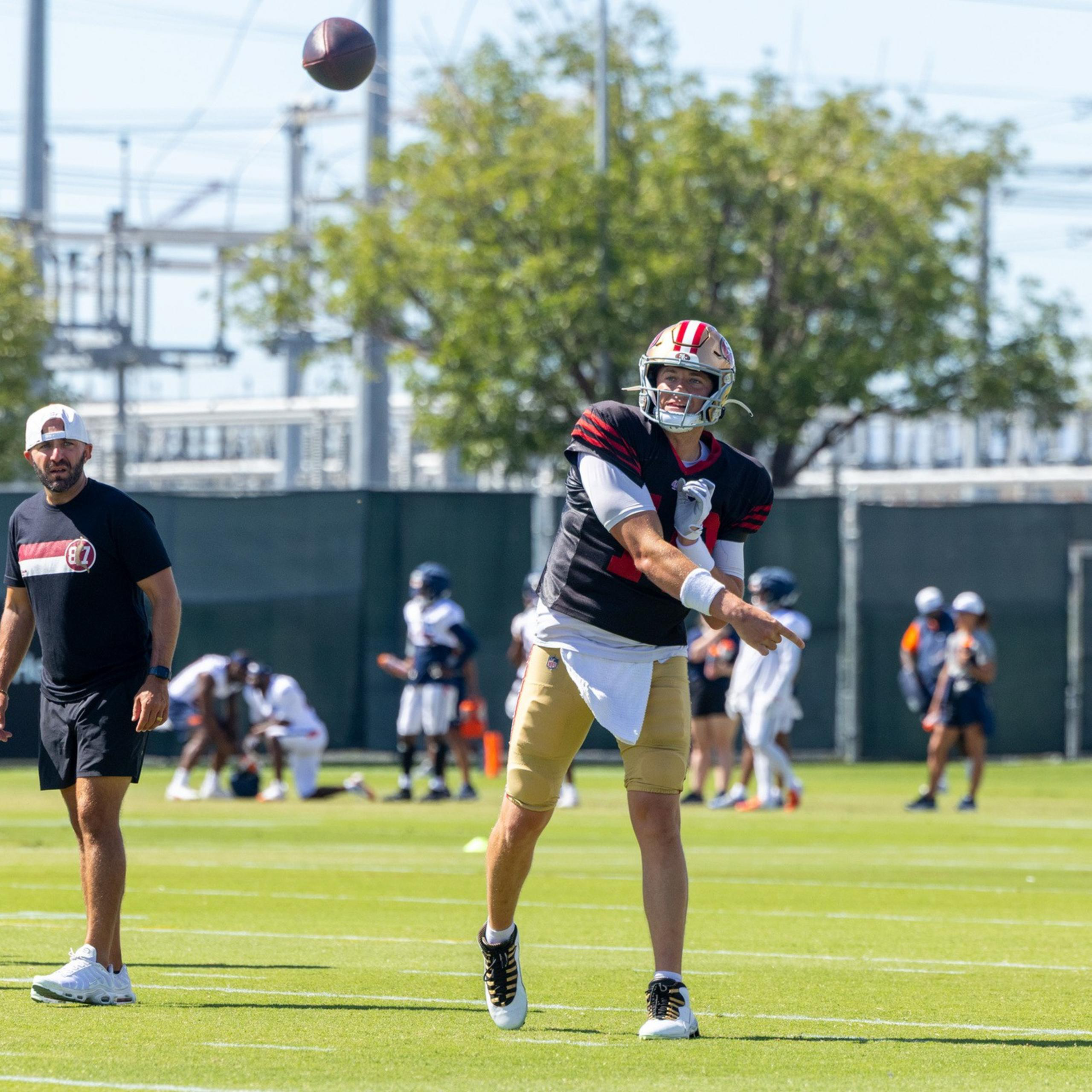 A football player in a black and gold uniform throws a football while coaches and other players stand nearby on a sunny practice field.
