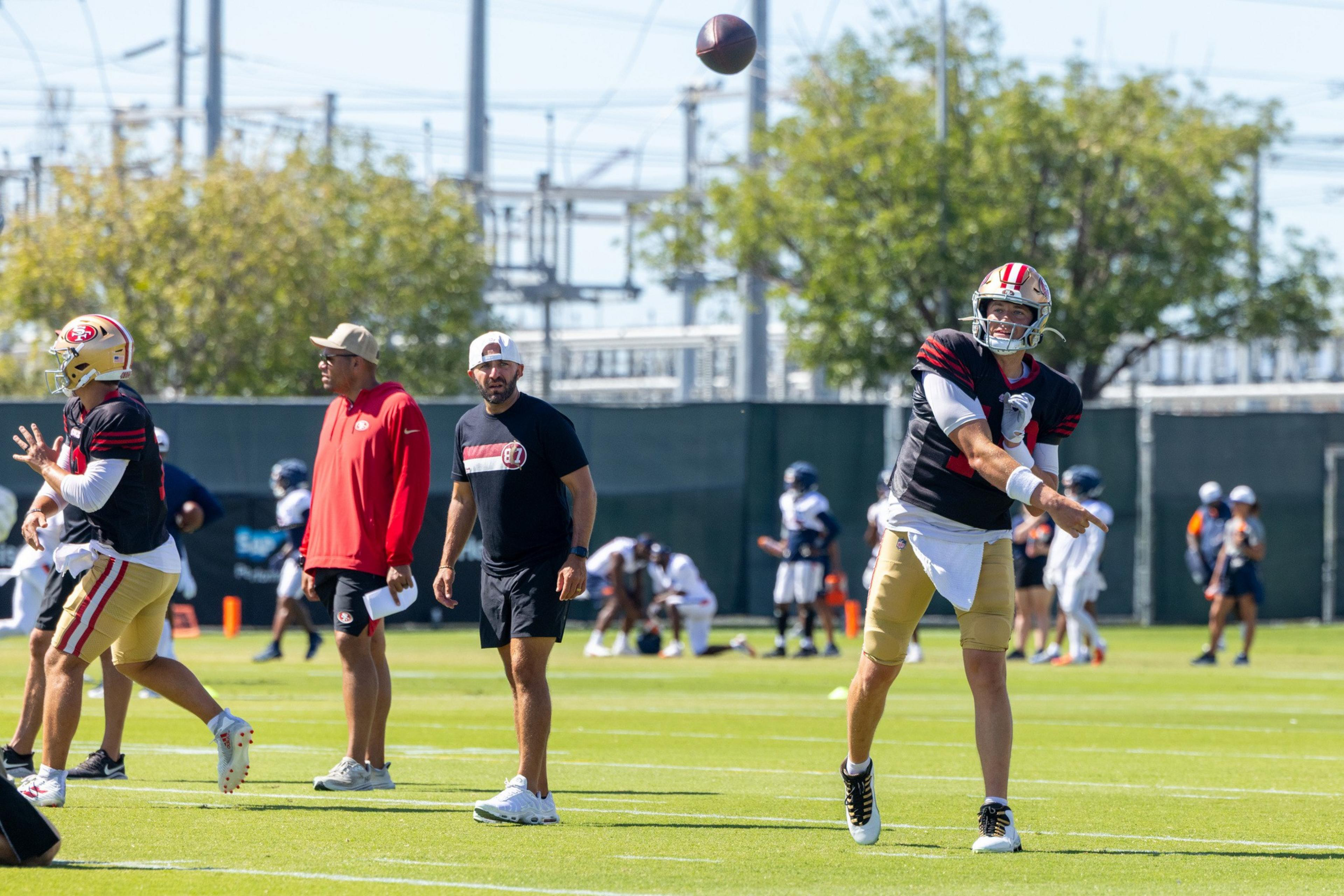 A football player in a black and gold uniform throws a football while coaches and other players stand nearby on a sunny practice field.