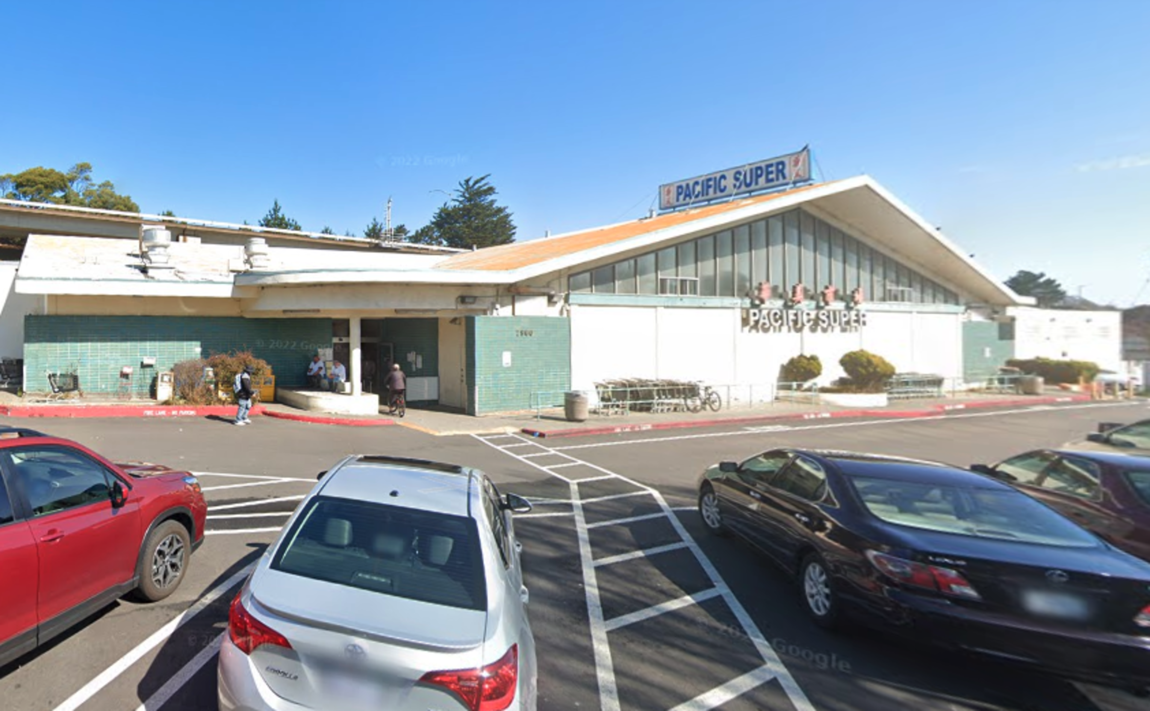 A one-story grocery store named "Pacific Super" with a triangular roof, green-tiled walls, and several parked cars in front under a clear blue sky.
