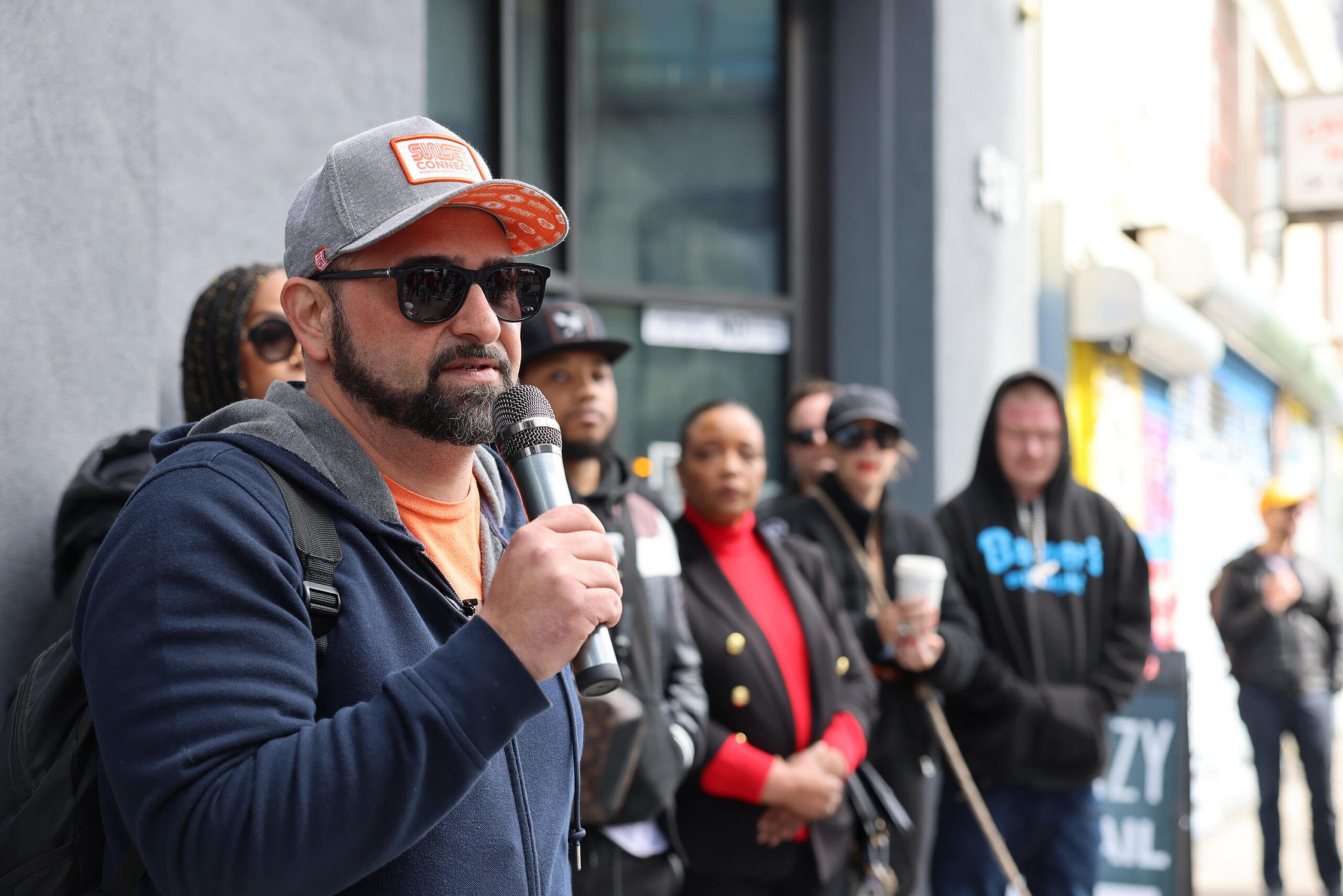 A bearded man in sunglasses and a cap speaks into a microphone while a diverse group of people listen behind him on a city sidewalk.
