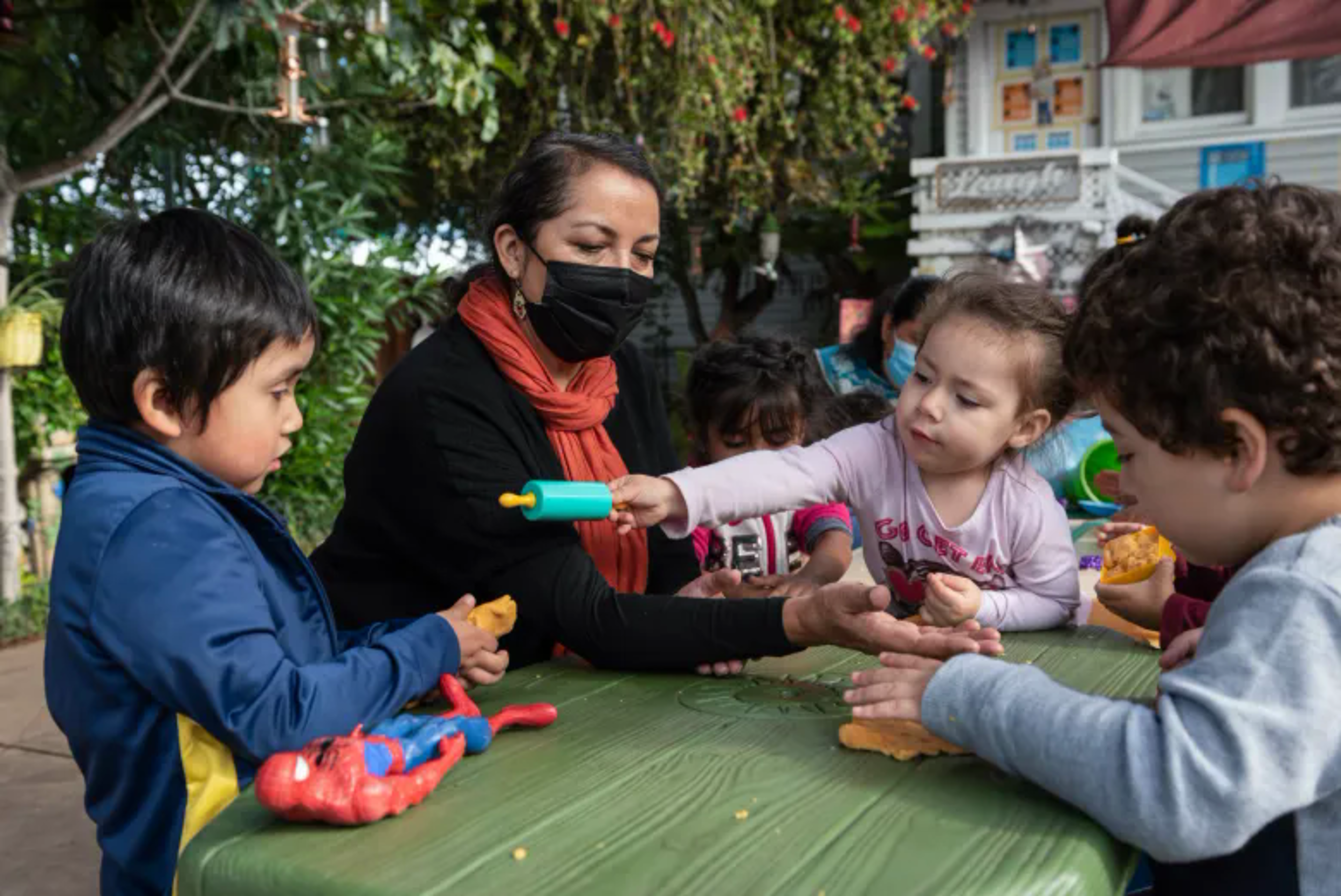 A woman wearing a mask helps four children playing with clay at a green table outdoors.