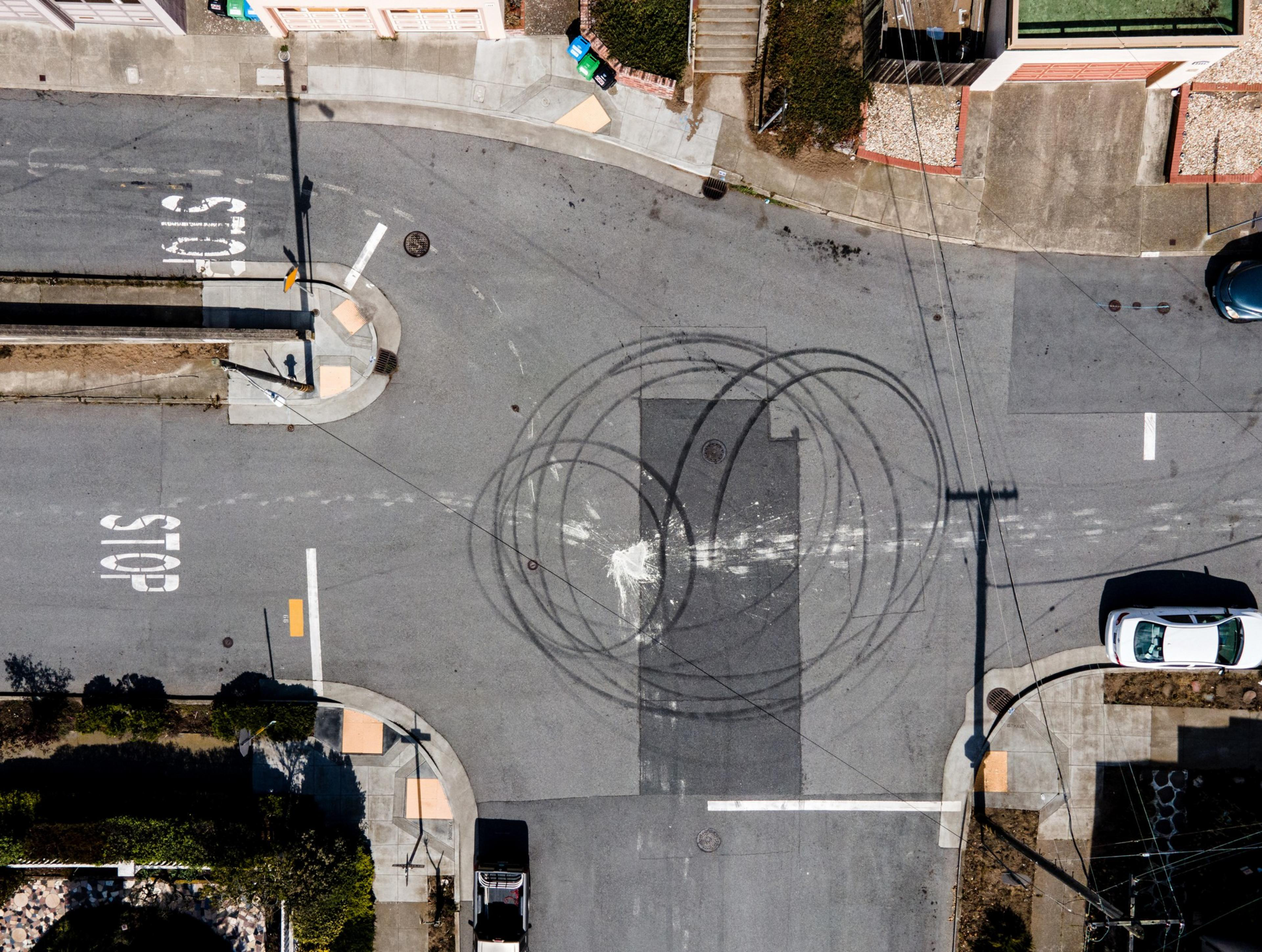 The image shows an aerial view of a street intersection with visible tire skid marks in circular patterns. There are &quot;STOP&quot; markings on the road and a white car.