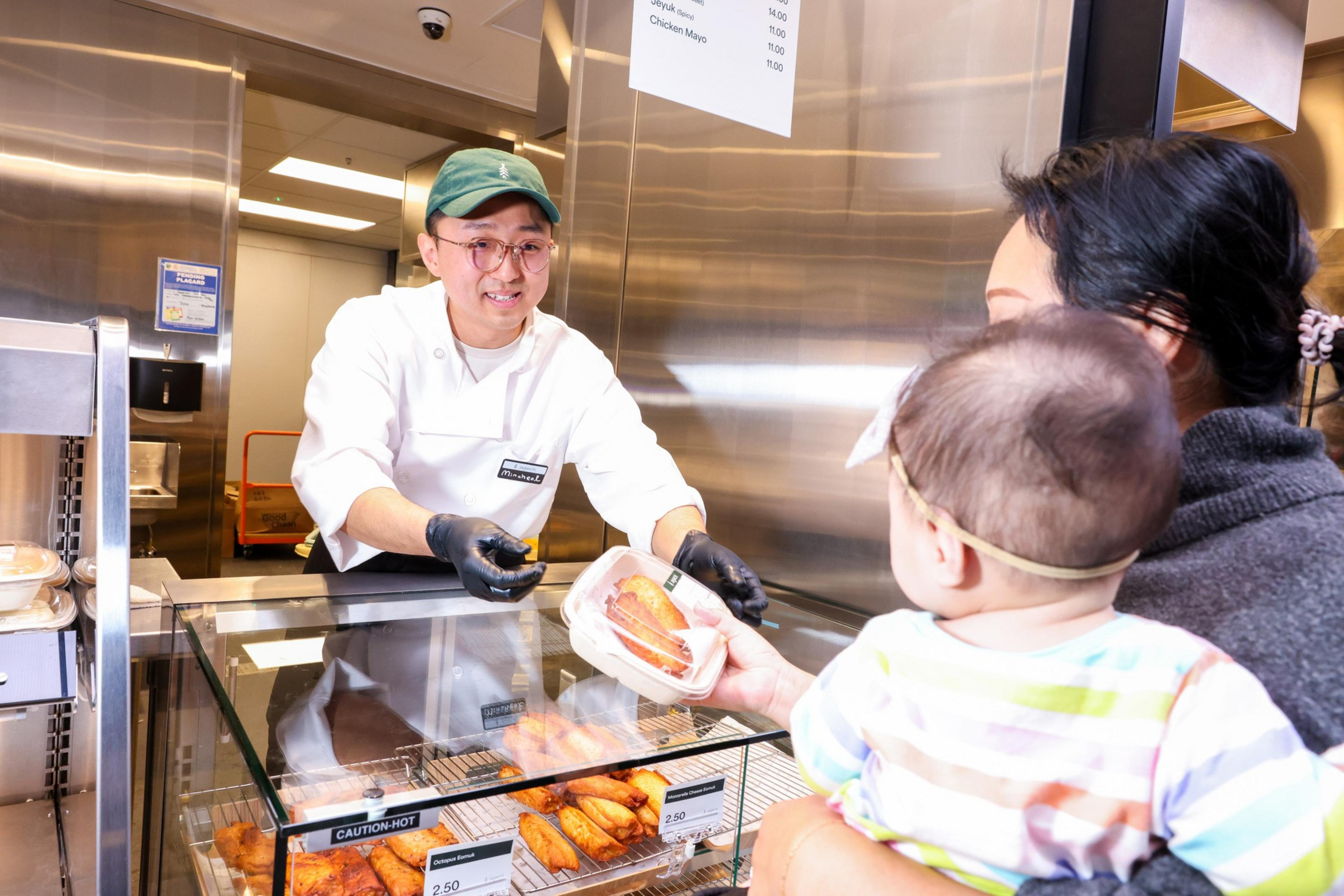 A friendly cashier, wearing a green cap and gloves, hands a packaged pastry to a woman holding a baby at a deli counter.
