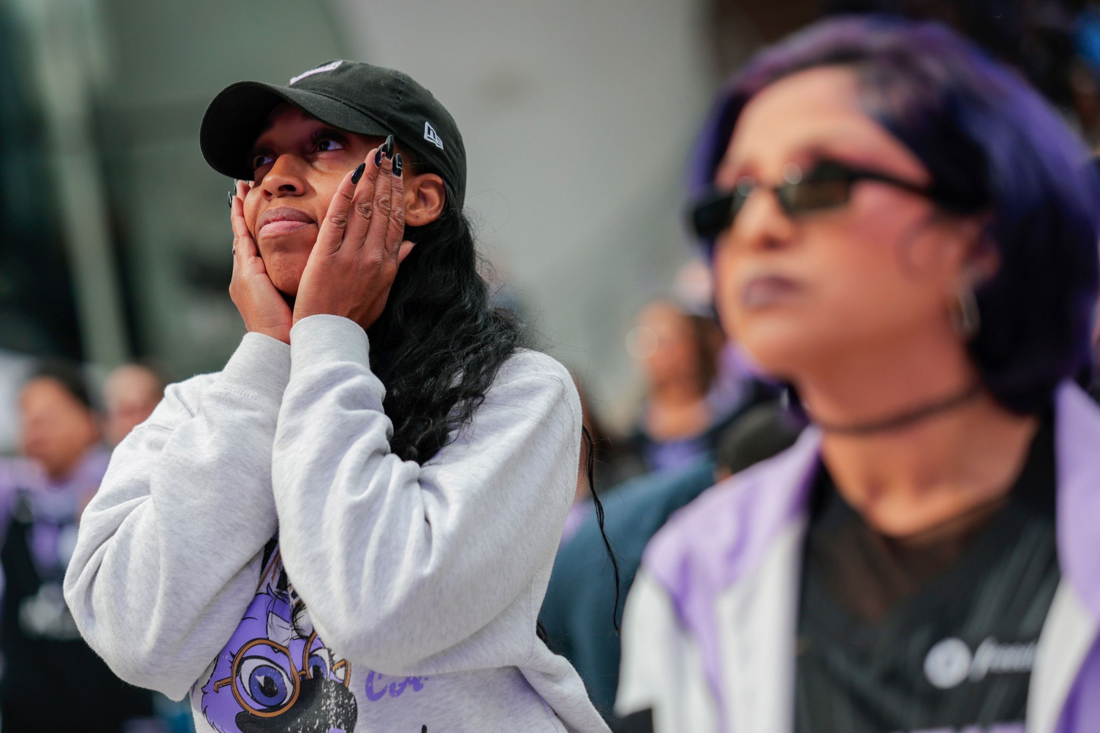 A person wearing a black cap and gray sweatshirt holds their face with both hands, looking upward with a thoughtful expression. Another person with purple hair 